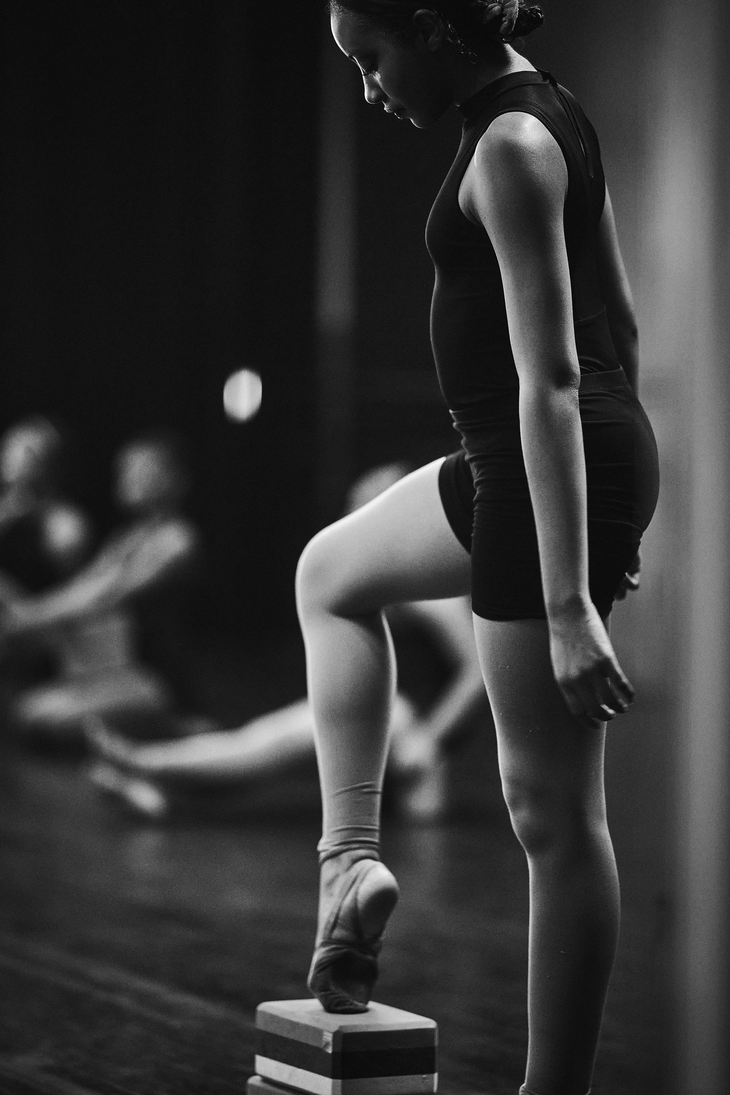 Black and white photo of a woman focusing during a weightlifting exercise, balancing on a small platform with her left foot, while her right leg is raised, and her hands are hanging down.