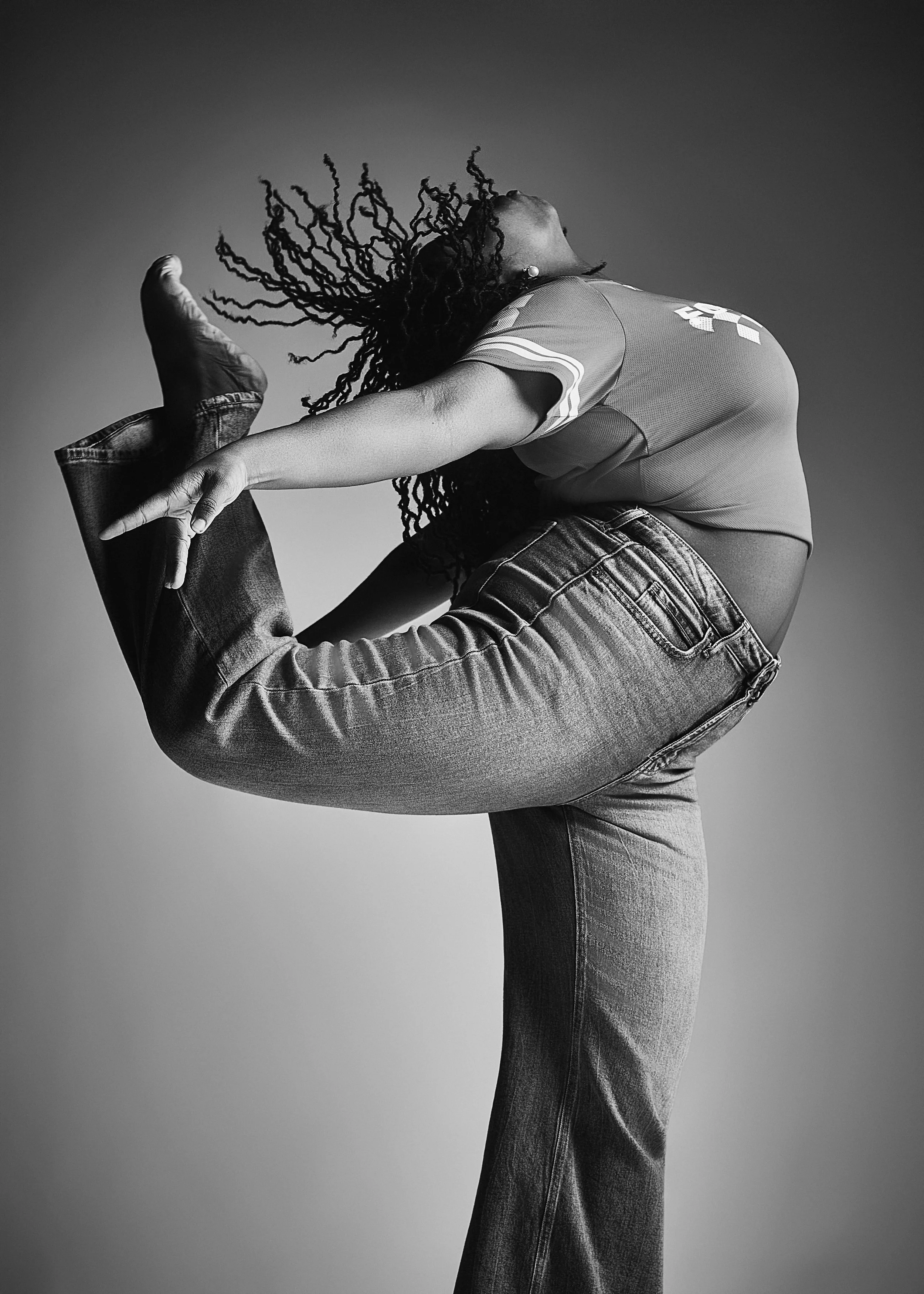 A person in a sports jersey and jeans is performing a yoga pose called Natarajasana or Lord of the Dance pose, with one leg raised behind and held by the hand, in a black-and-white studio photo.