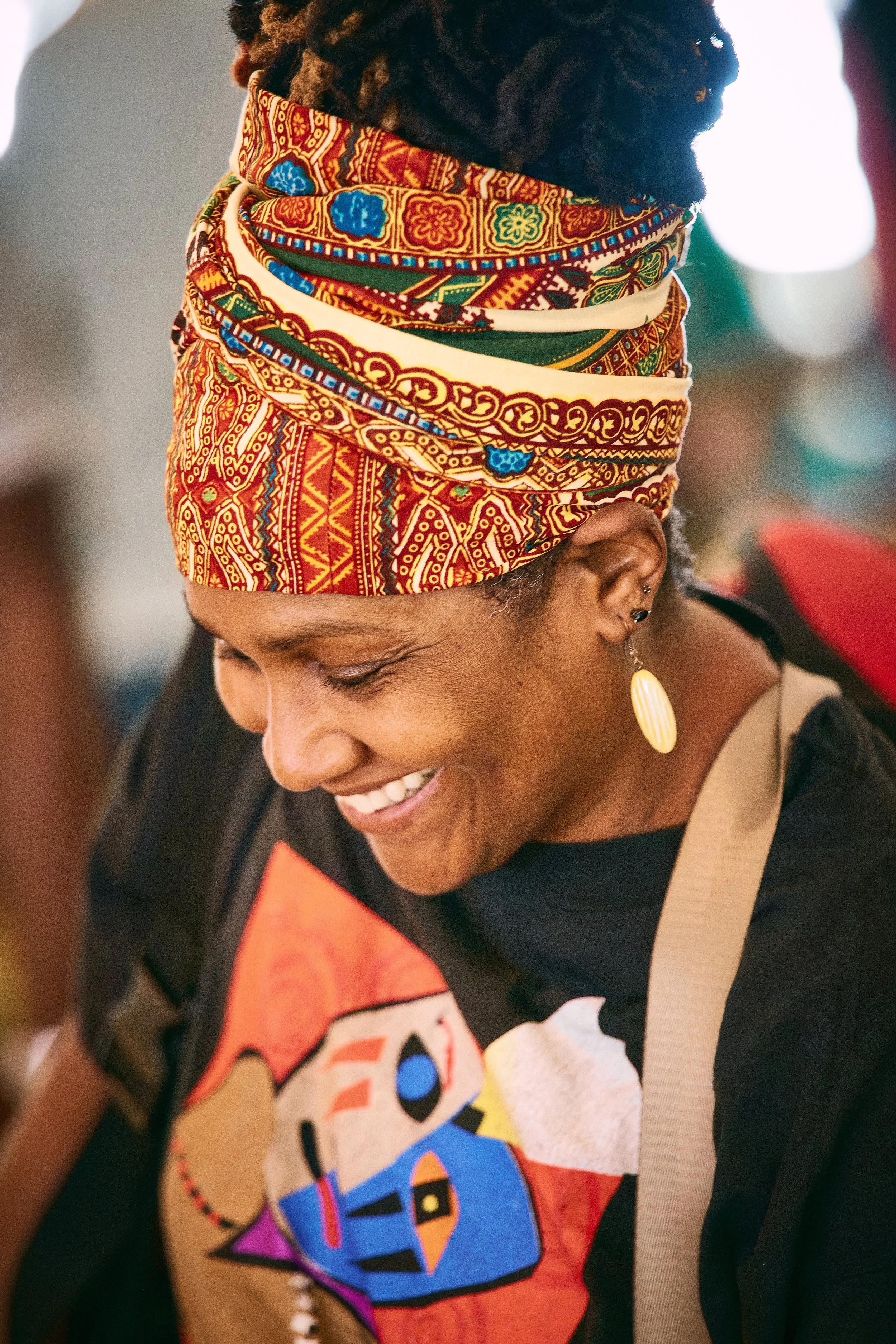 A woman wearing a colorful head scarf with red, orange, green, and blue patterns. She has a smile and is wearing earrings, a black shirt with a graphic print, and a beige strap across her shoulder.