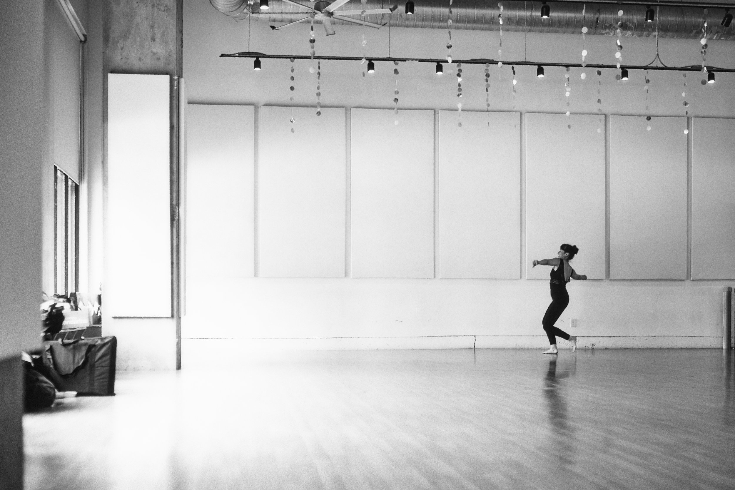 A dancer practicing in a spacious dance studio with large windows, white wall panels, and hanging decorations from the ceiling. The dancer is mid-movement with arms extended and legs bent, wearing dark athletic clothing and shoes.