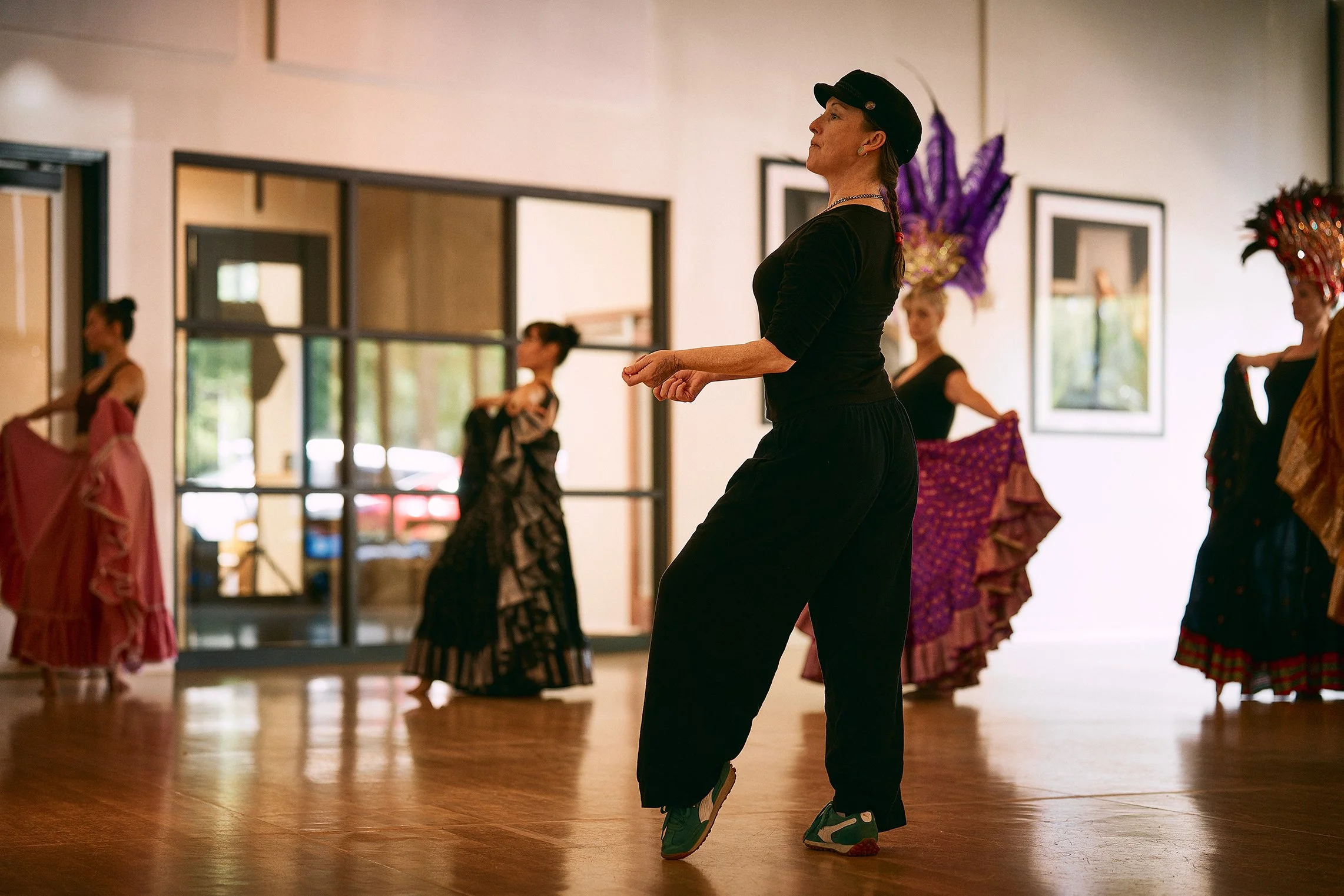 Women participating in a traditional dance class, wearing colorful costumes, in a well-lit studio with wooden floors and framed pictures on the wall.