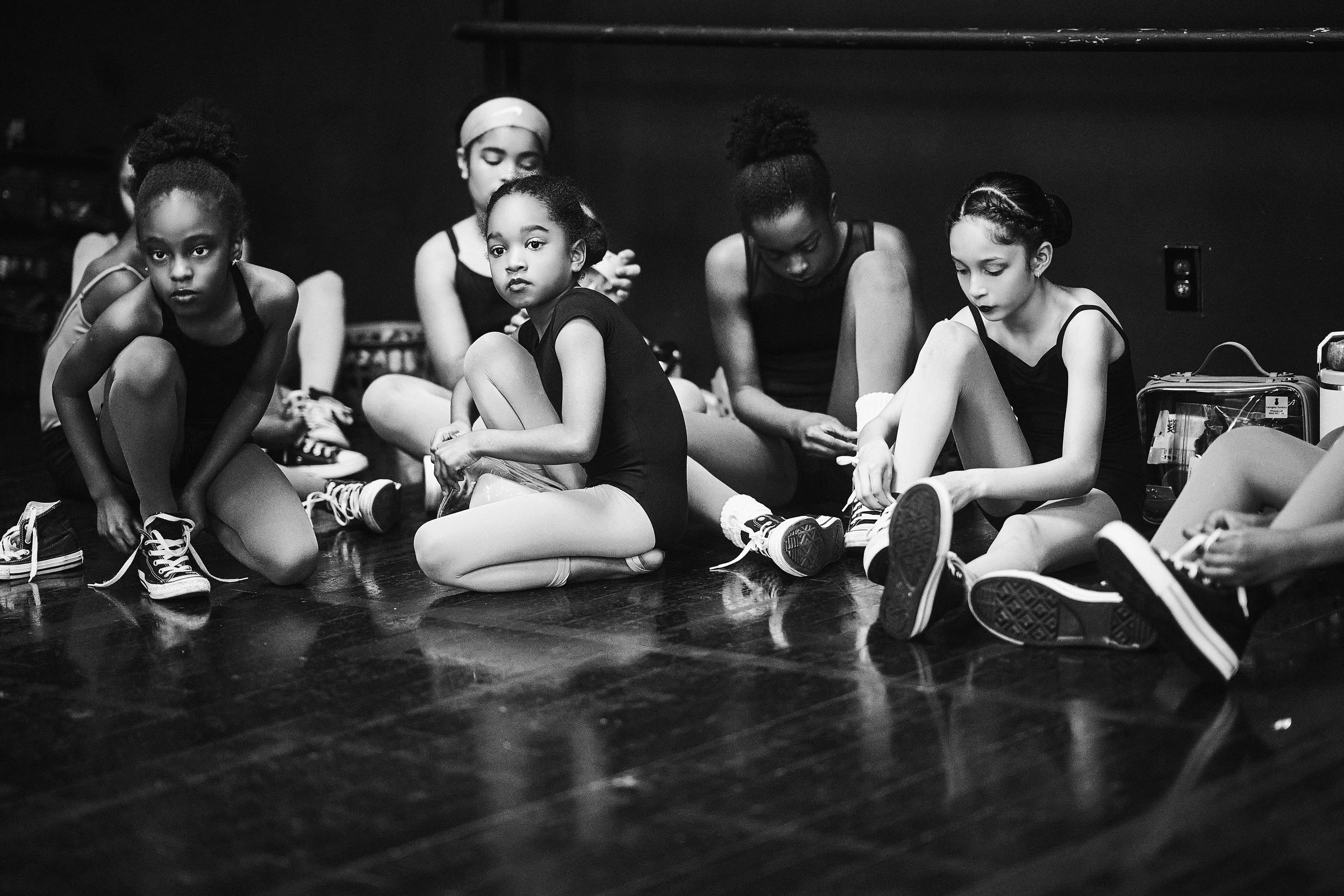 A group of young girls sitting on the floor, tying their shoelaces, in a dance studio with a dark wall and floor.