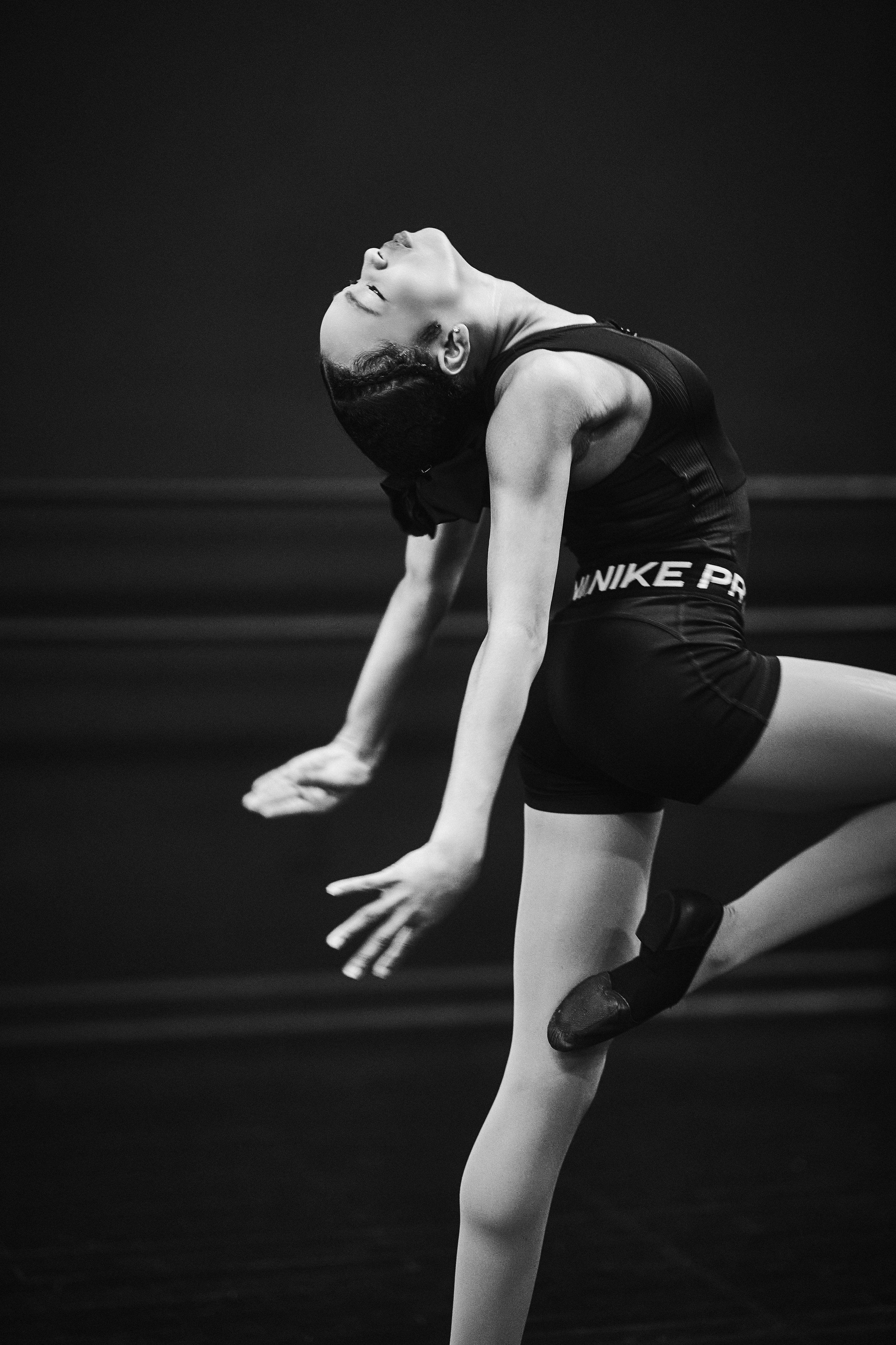 A female ballet dancer in a black sports outfit, performing a flexible pose on stage with her back arched and head tilted back.