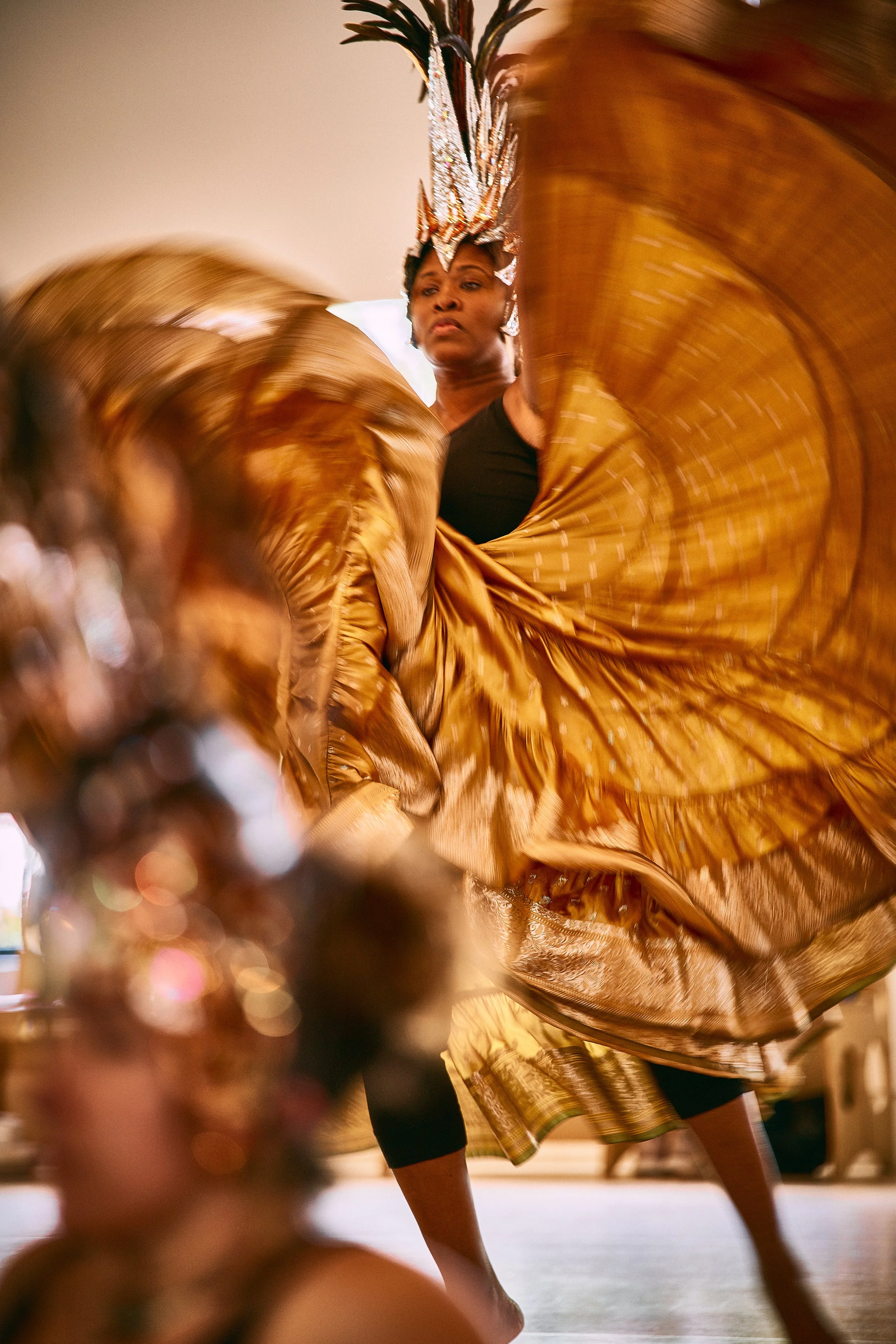 A dancer wearing an elaborate golden costume with flowing fabric, a headpiece with feathers, and black leg coverings performing on stage.