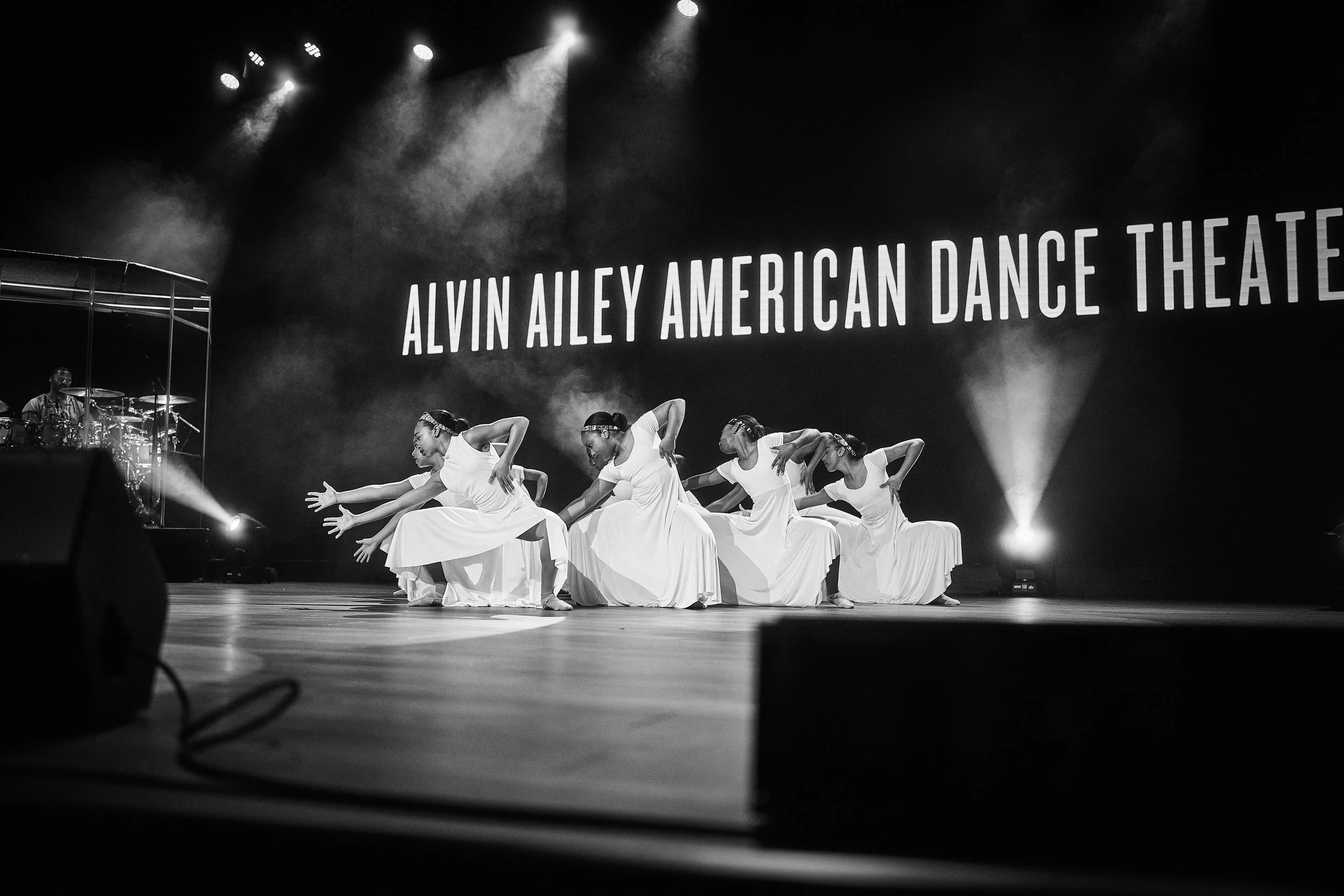 A dance performance on stage at the Alvin Ailey American Dance Theatre. Dancers in white costumes are kneeling with arms extended, with stage lights and a large illuminated sign in the background.