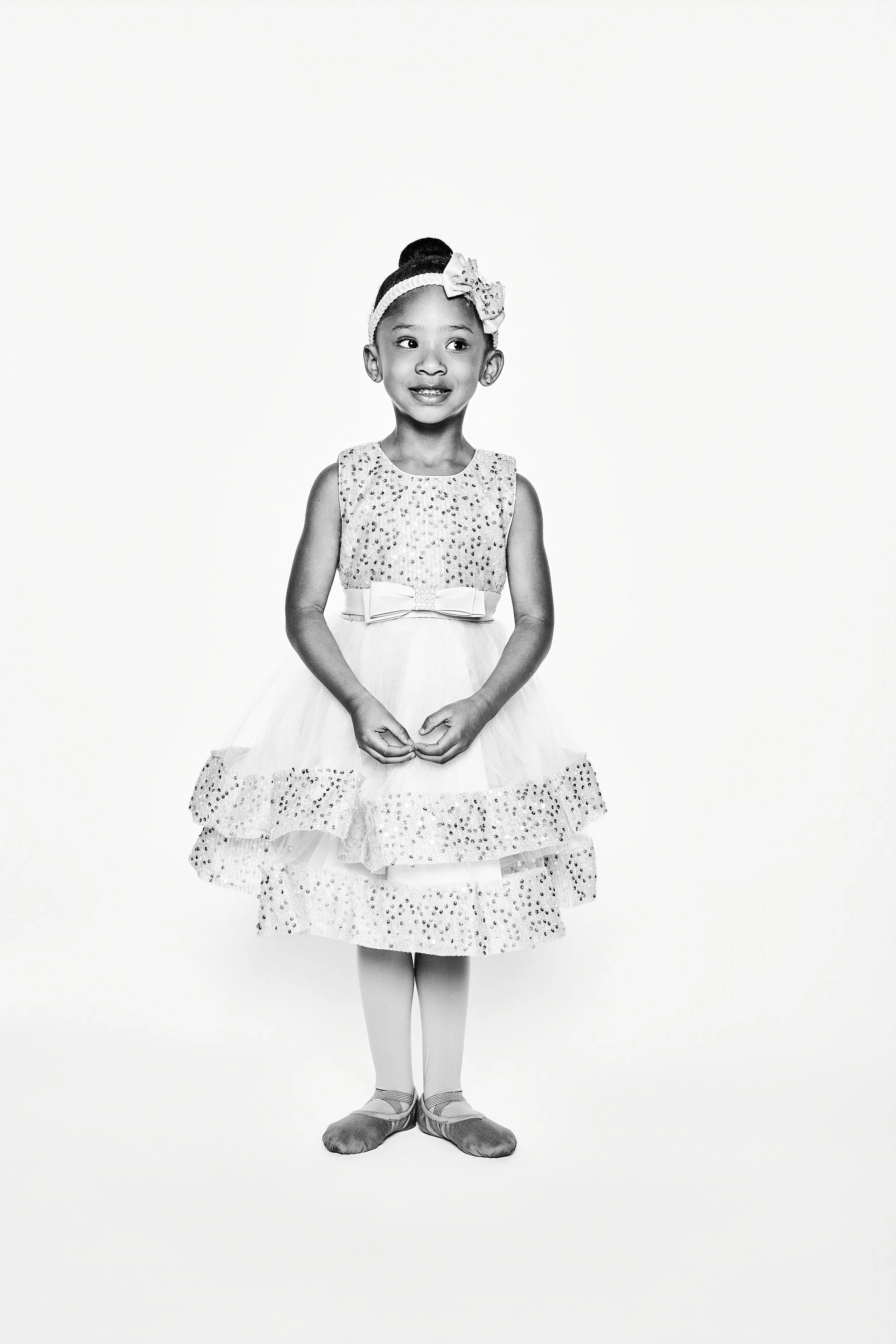 A young girl in a princess or ballerina costume, standing against a plain background, looking to her left with a slight smile, wearing a dress with sequins, a bow on her waist, and a headband with a bow, with her hands clasped in front of her.