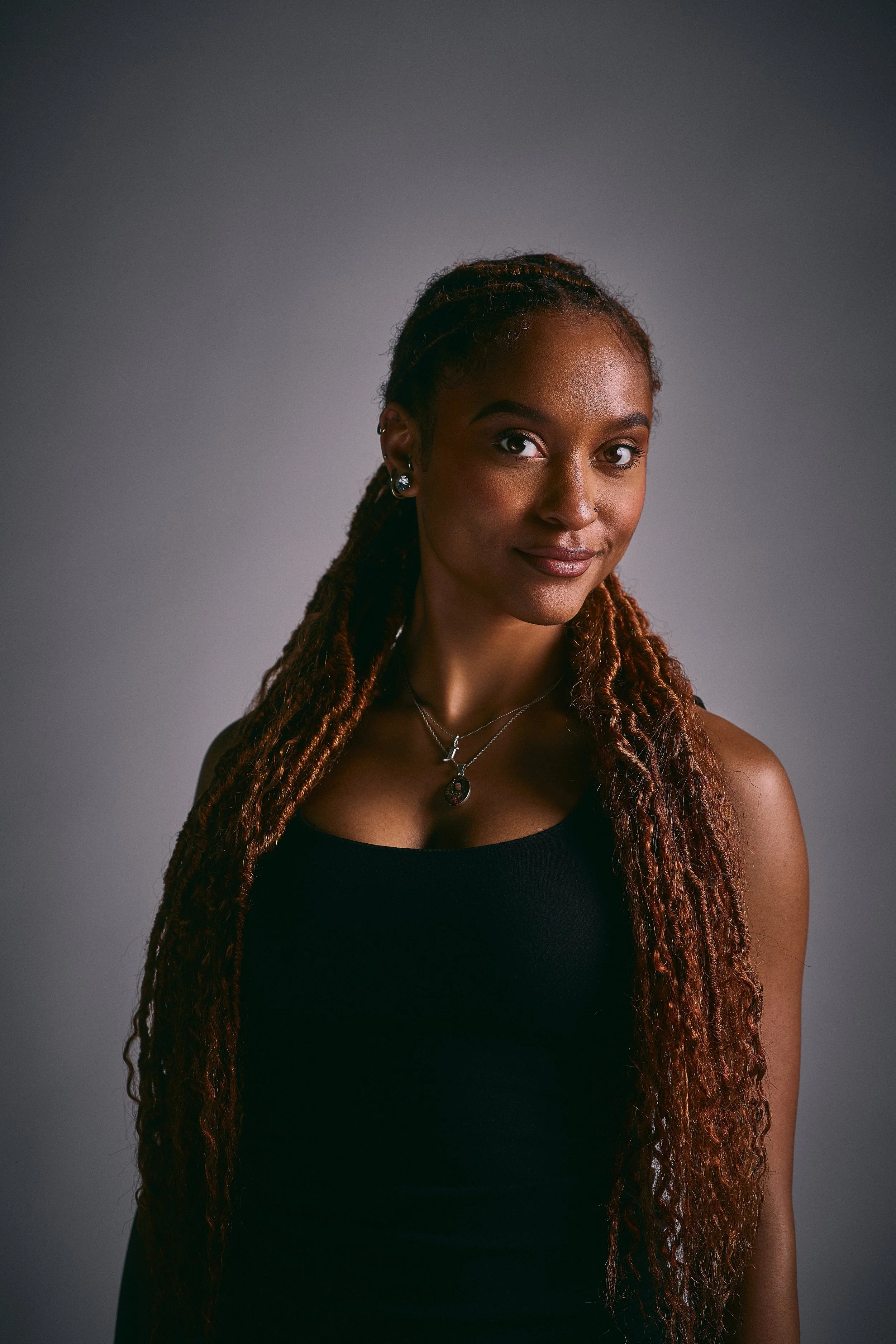 A portrait of a young African American woman with long braided hair, wearing a black sleeveless top, earrings, and layered necklaces, standing against a gray background.