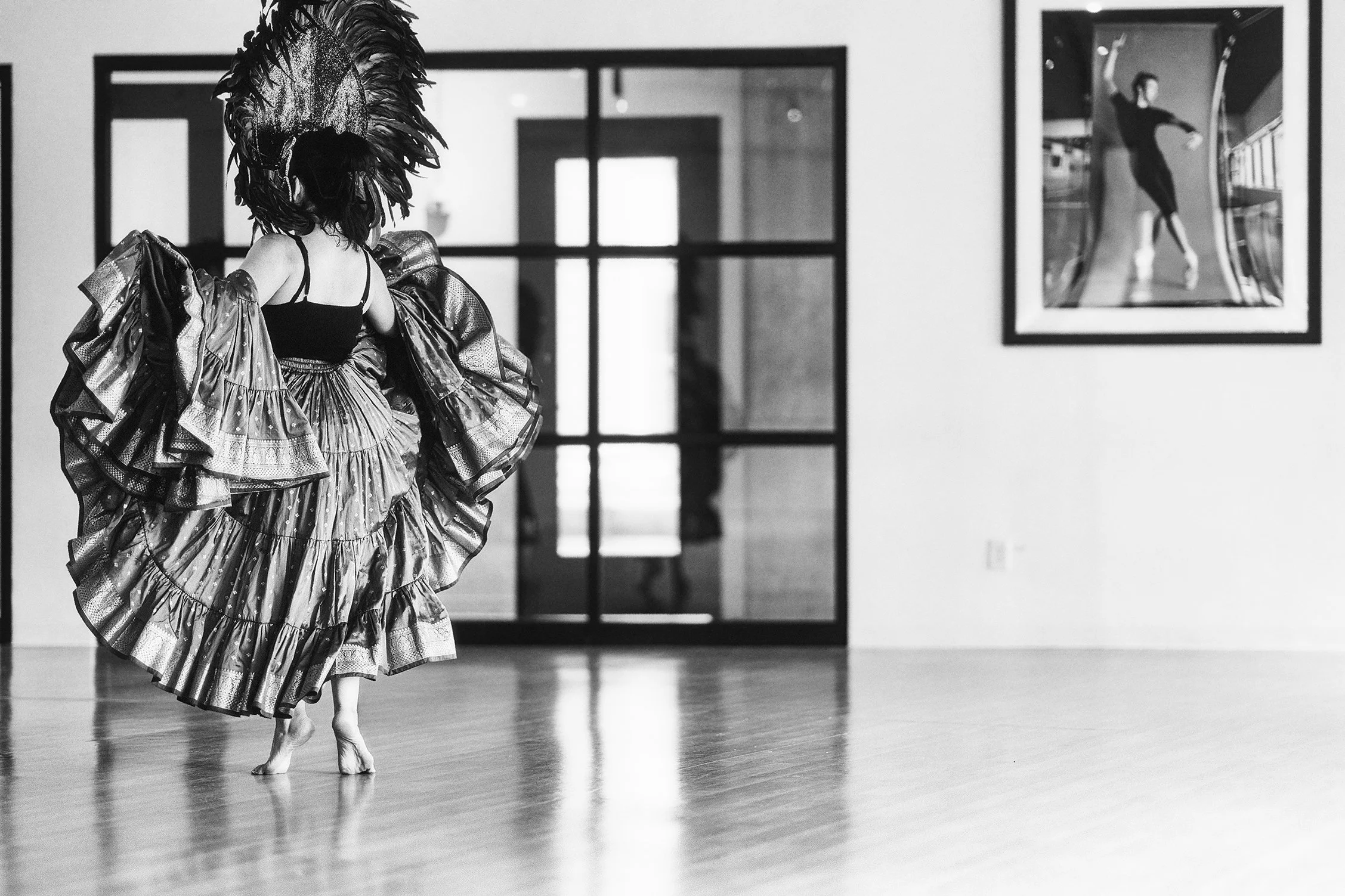 A ballet dancer wearing a costume with a large ruffled skirt and an elaborate feathered headpiece practices in a dance studio, with framed artwork on the wall in the background.