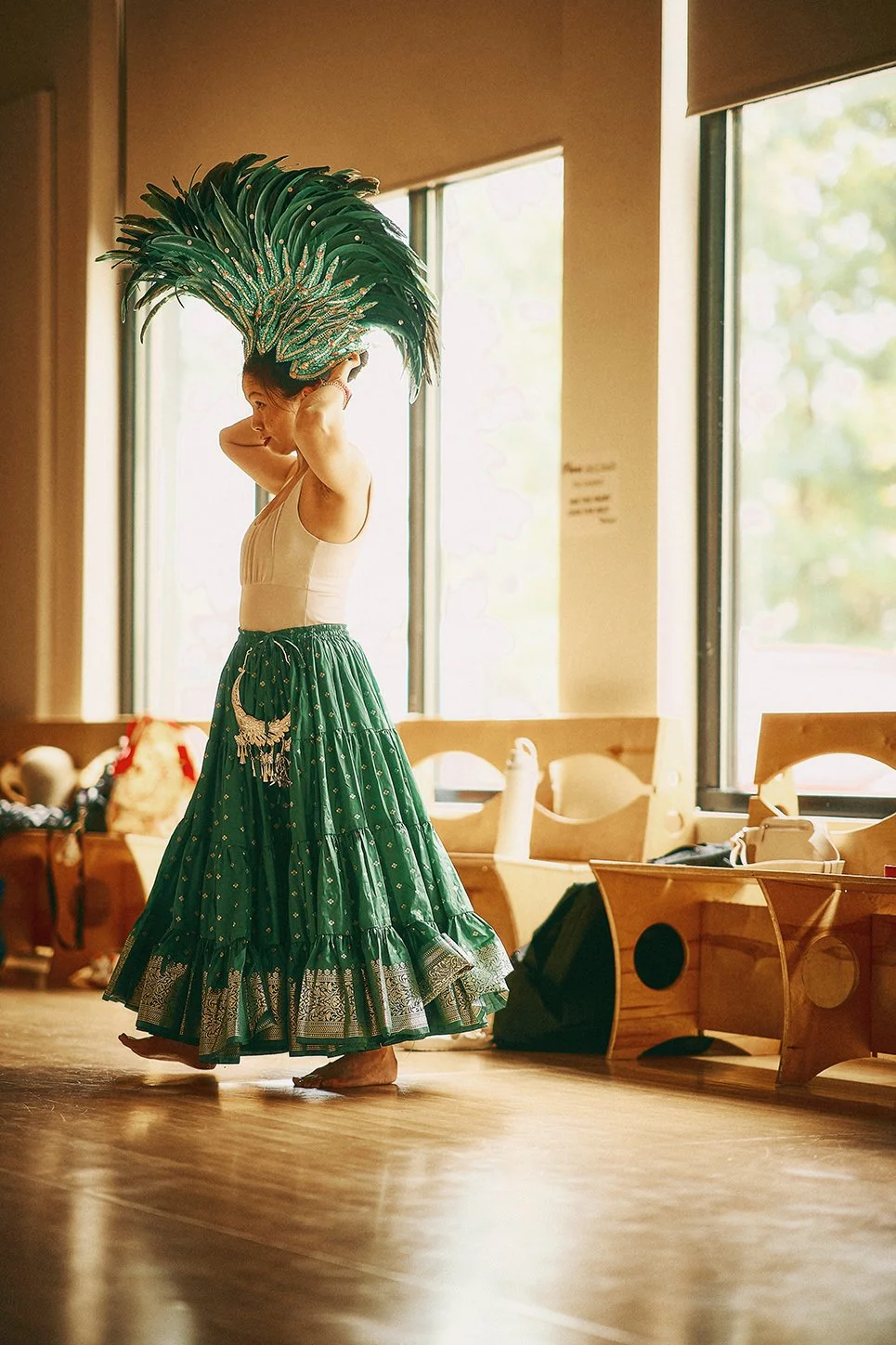 A woman dressed in a traditional Indian dance costume with a green skirt and large feathered headpiece, practicing a dance in a sunlit room with wooden floors and large windows.