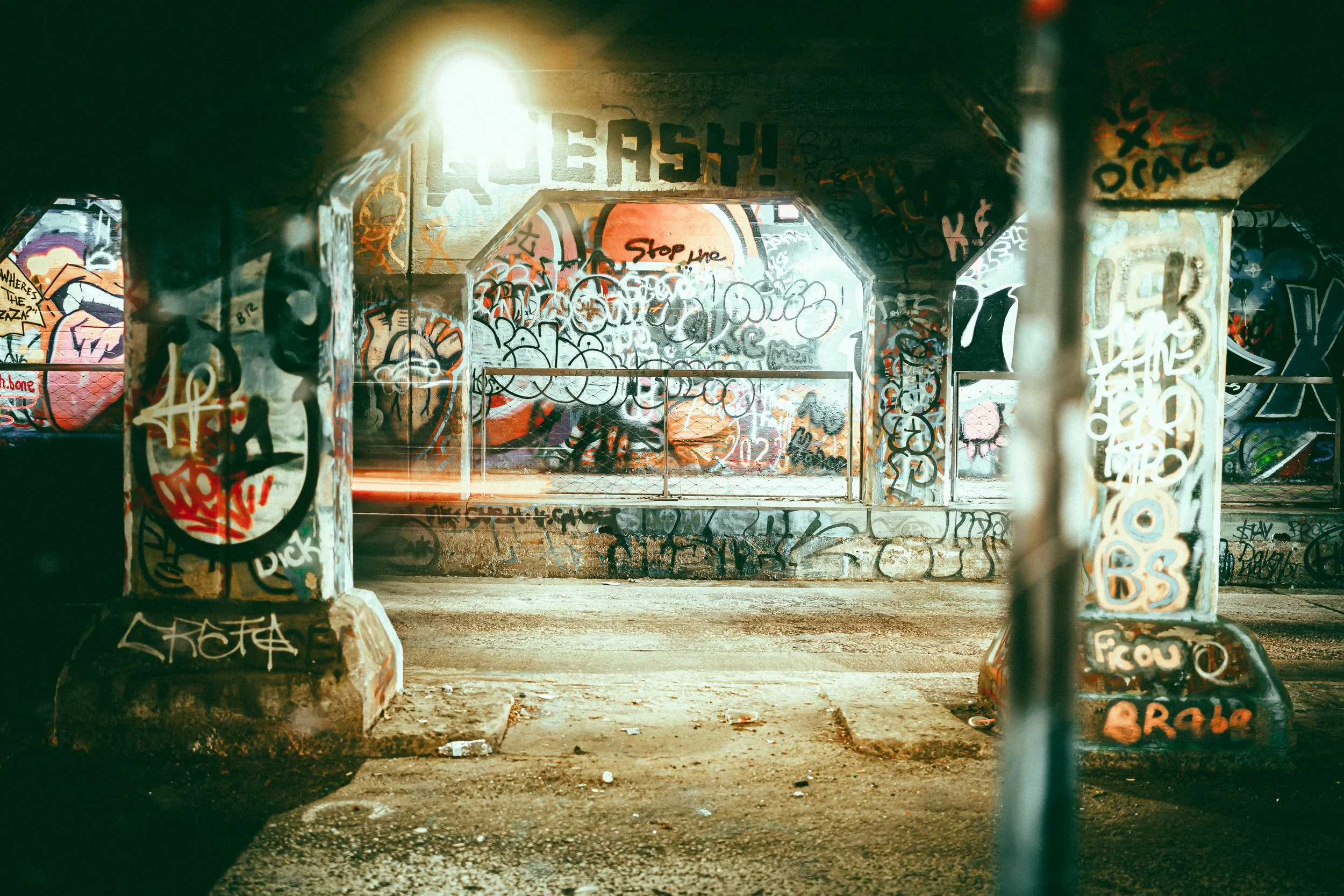 Graffiti-covered urban underpass with columns and walls, dimly lit.