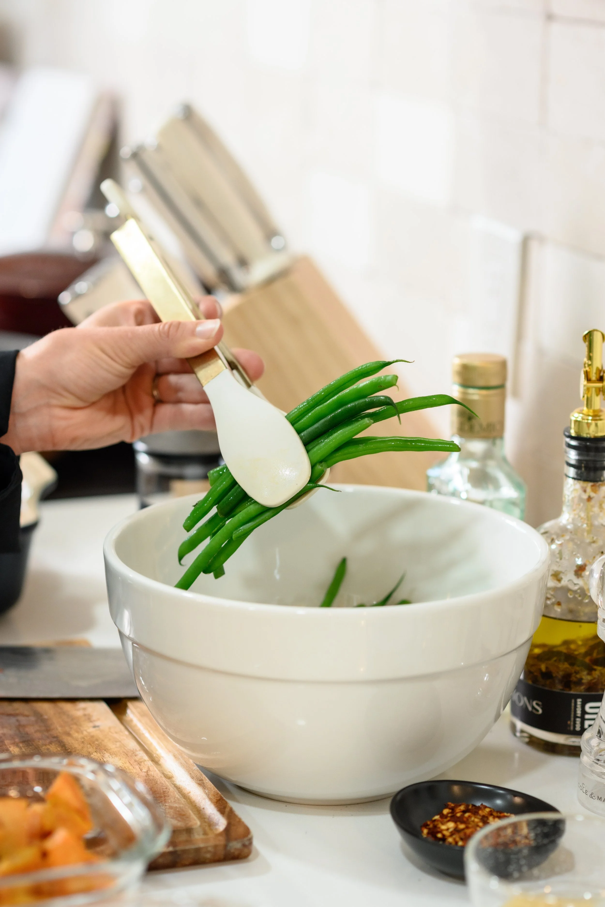 Person rinsing green beans in a white bowl, using a white and gold spatula, with kitchen items in the background.