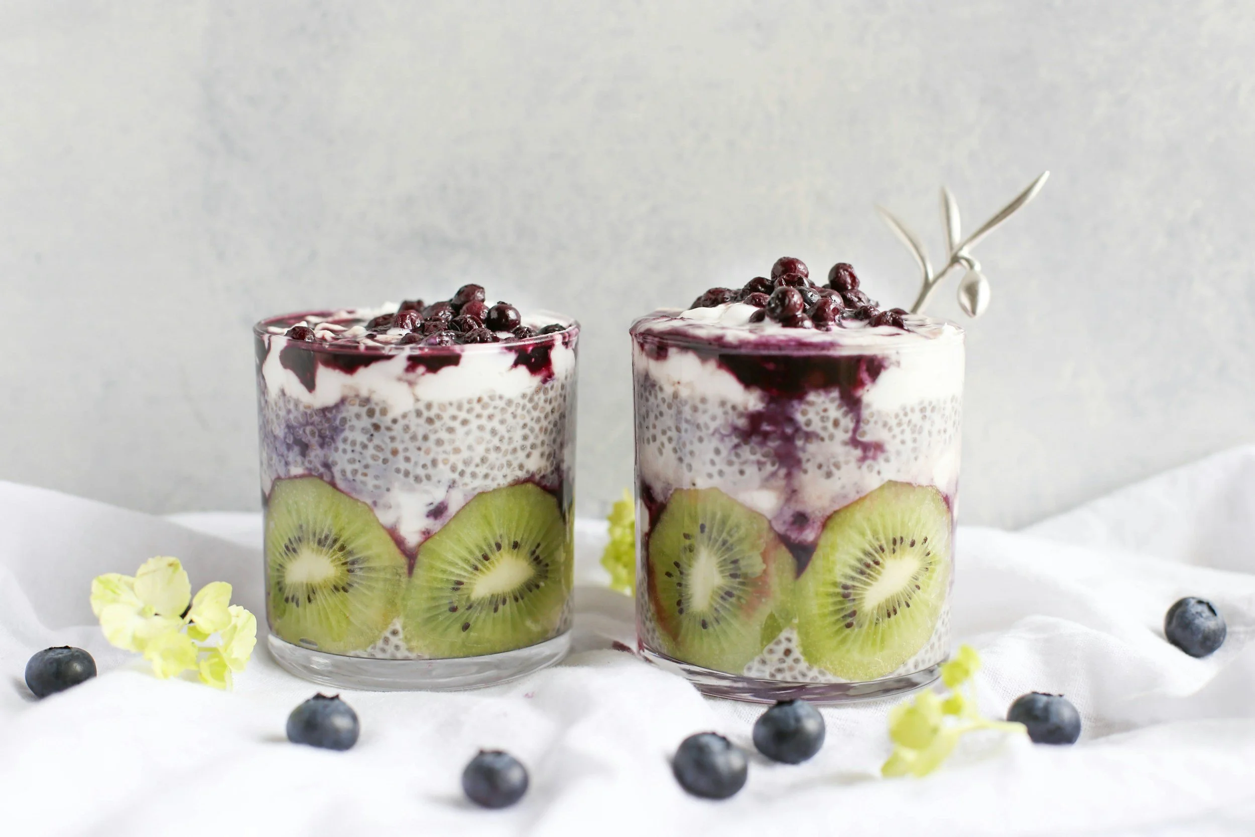 Two glasses of layered fruit and chia seed pudding with kiwi slices, blueberries, and berry compote, on a white surface with scattered blueberries and flowers.