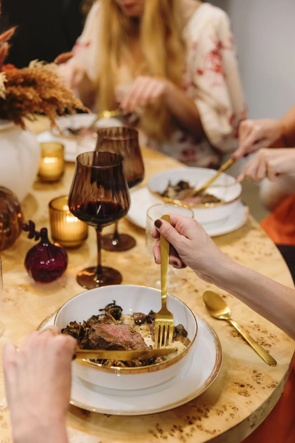 People gathered around a dinner table with glasses of red wine, eating a meal with a variety of dishes, including a bowl with a piece of steak and vegetables, and decorated with candleholders and vases.