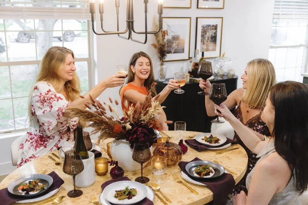 Five women celebrating and raising glasses of wine and drinks at a dinner party around a decorated table in a well-lit room.