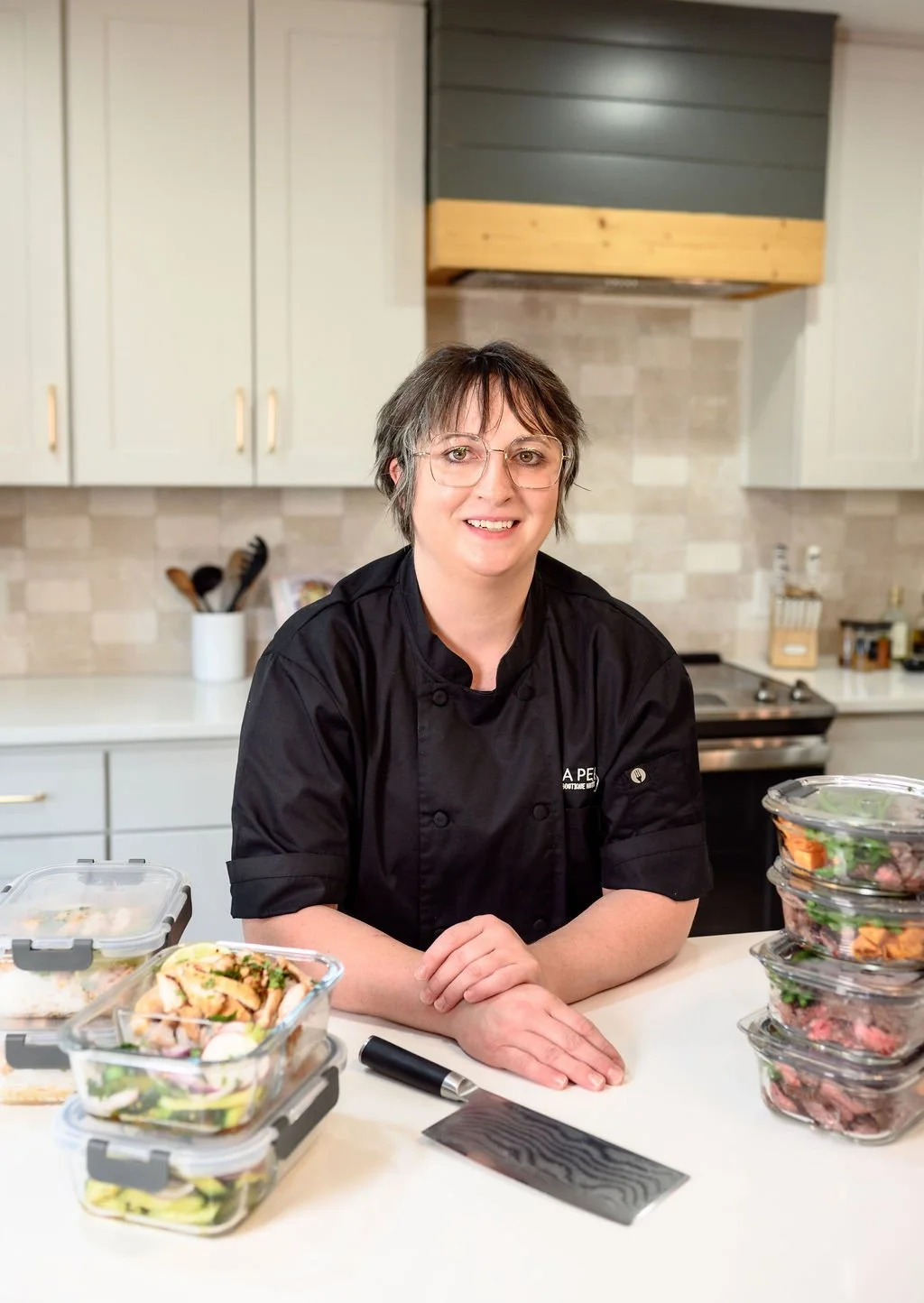 A woman dressed in a black chef's coat sitting at a kitchen counter with prepared salads in plastic containers, smiling at the camera.