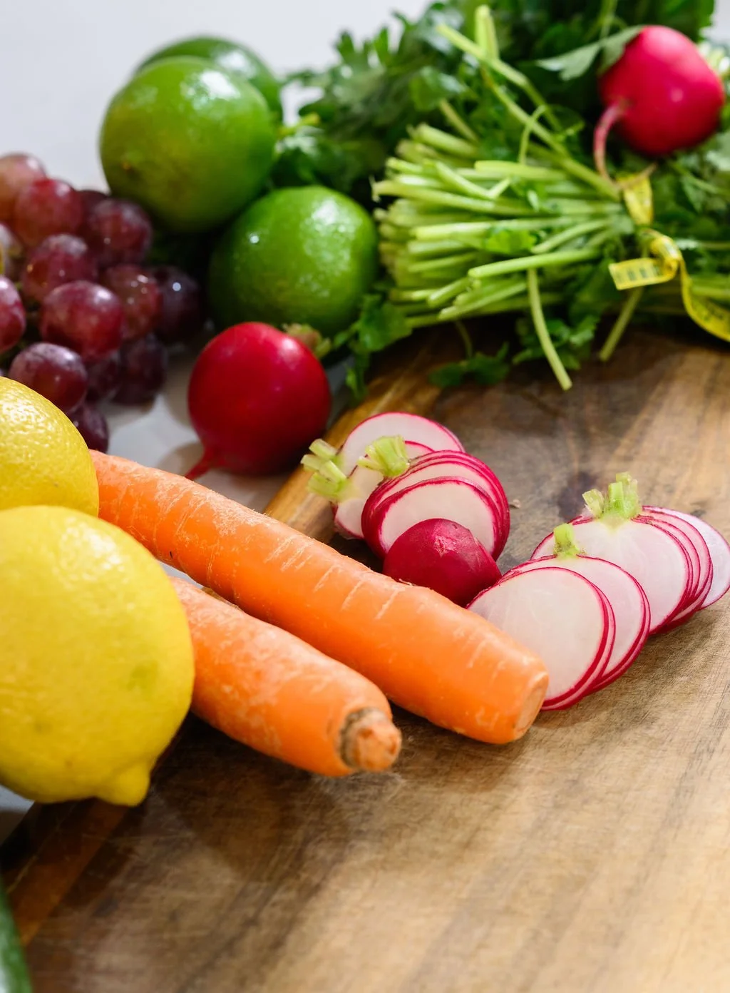 Fresh vegetables and fruits including carrots, radishes, lemons, grapes, and limes on a wooden cutting board.