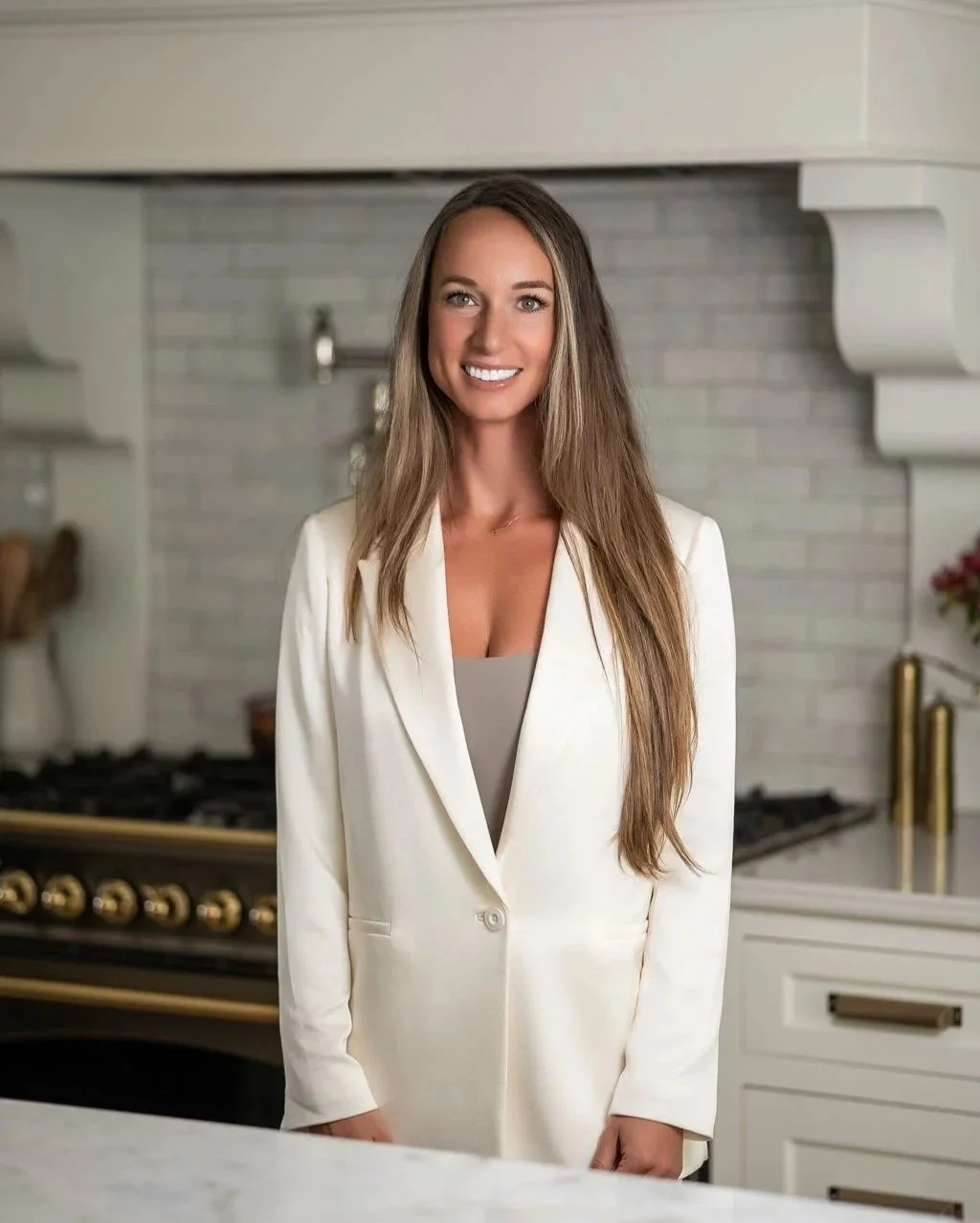 A woman with long brown hair wearing a cream-colored blazer and a beige top standing in a modern kitchen with white cabinets and a stainless steel stove.