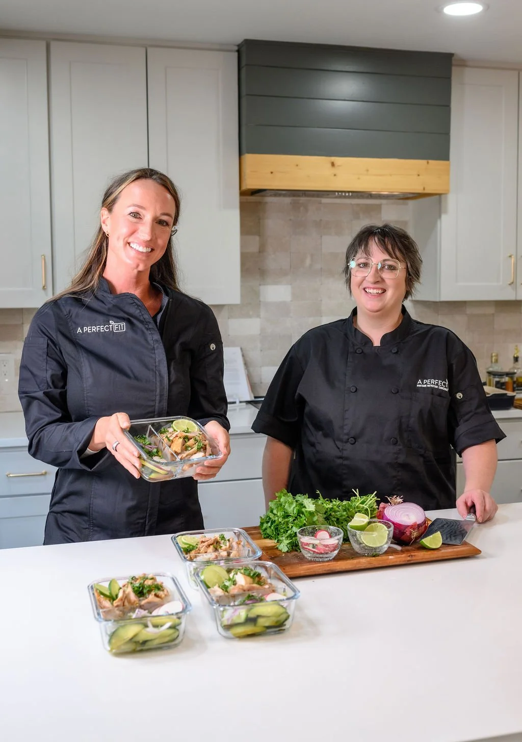 Two women in black chef jackets standing in a kitchen, preparing and showcasing bowls of a colorful dish with fresh herbs, vegetables, and lime slices.
