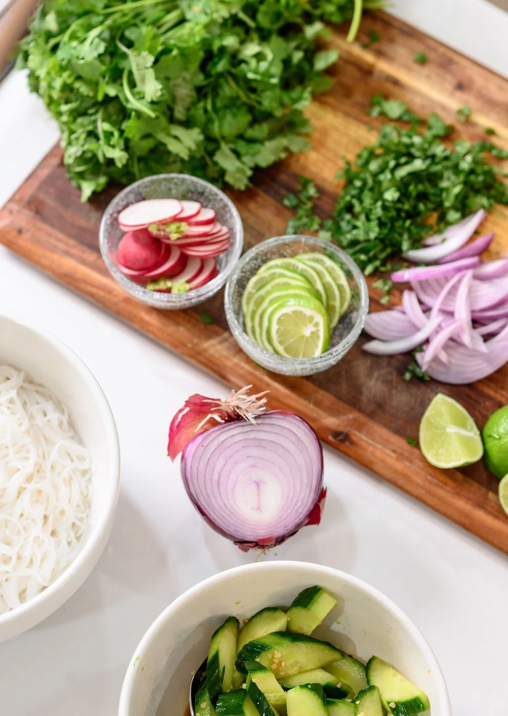Fresh herbs, radishes, limes, and sliced onions on a wooden cutting board, with sliced cucumbers in a white bowl, and a purple onion and noodles in separate white bowls nearby.