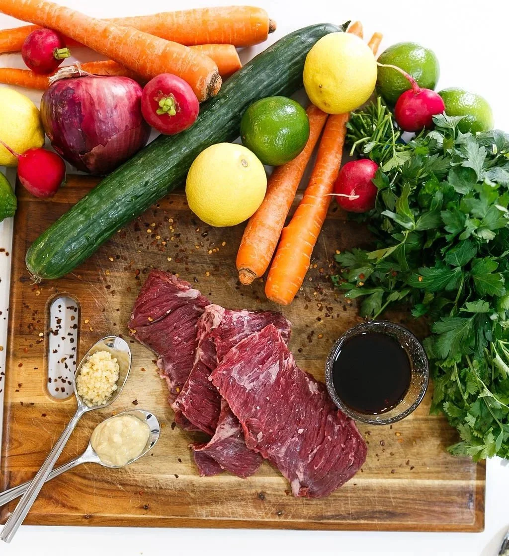 Fresh vegetables, including carrots, radishes, cucumbers, lemons, and green herbs, alongside raw beef and condiments on a wooden cutting board.