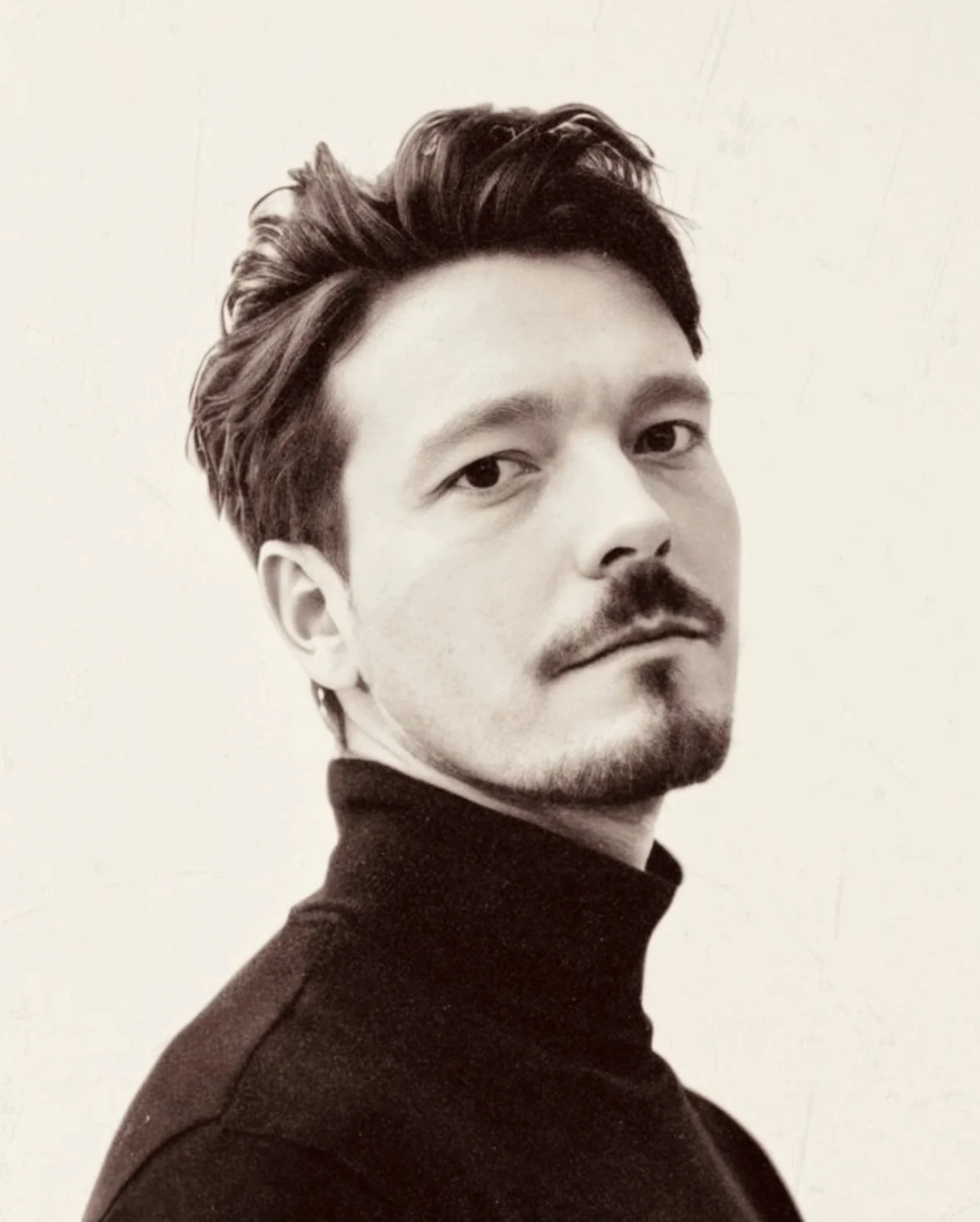 Black and white portrait of a man with short hair and a beard, wearing a turtleneck, looking at the camera.