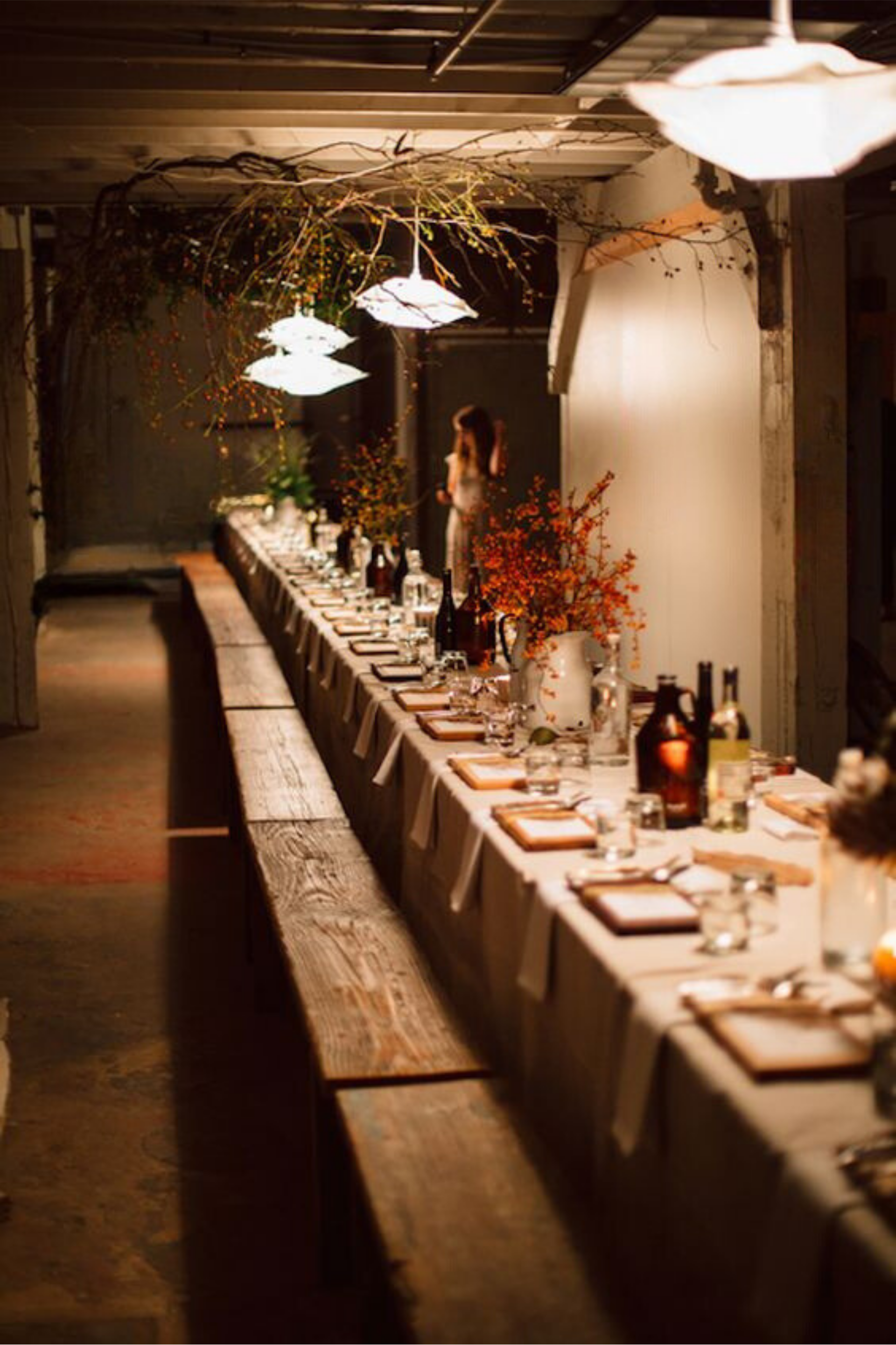 A long dining table set for a meal with white tablecloth, cutlery, glasses, and small bottles. Decorated with tall vases of orange and yellow flowers and branch arrangements hanging from the ceiling. Lighting fixtures hang above the table, creating a warm atmosphere in a rustic, industrial-style room.