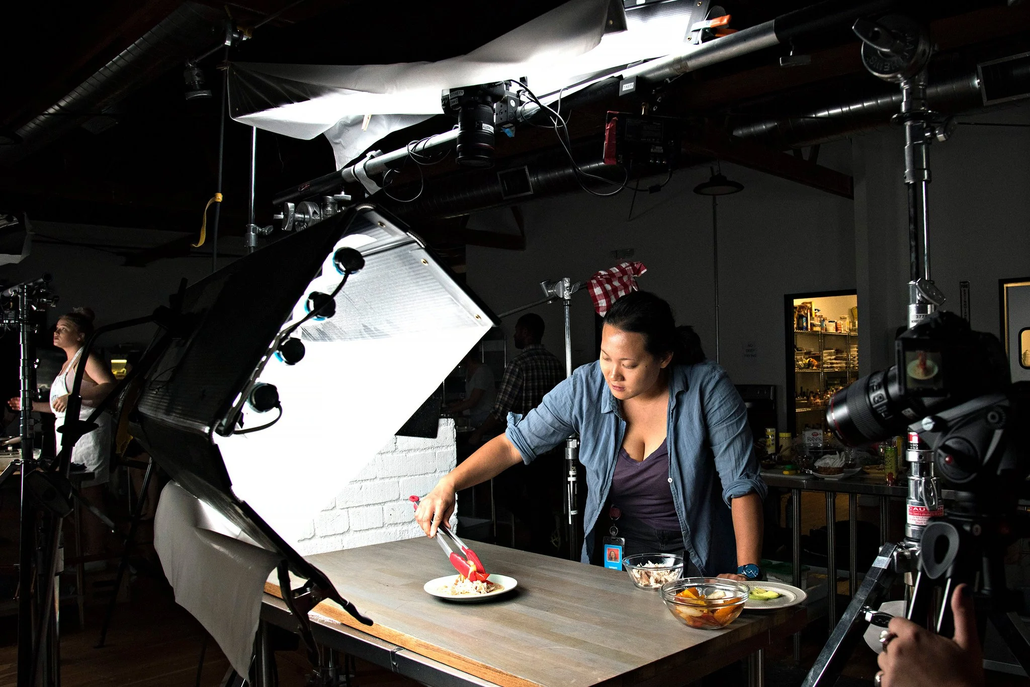 A person working on food presentation at a table in a professional kitchen, surrounded by photographic and lighting equipment.