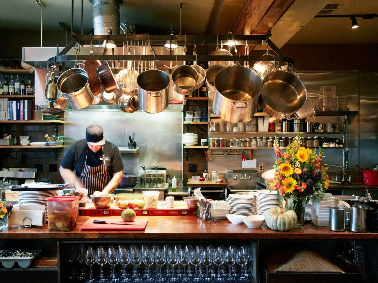 A chef preparing food in a professional kitchen with hanging pots and pans, a bouquet of flowers, and various kitchen supplies on the counter.