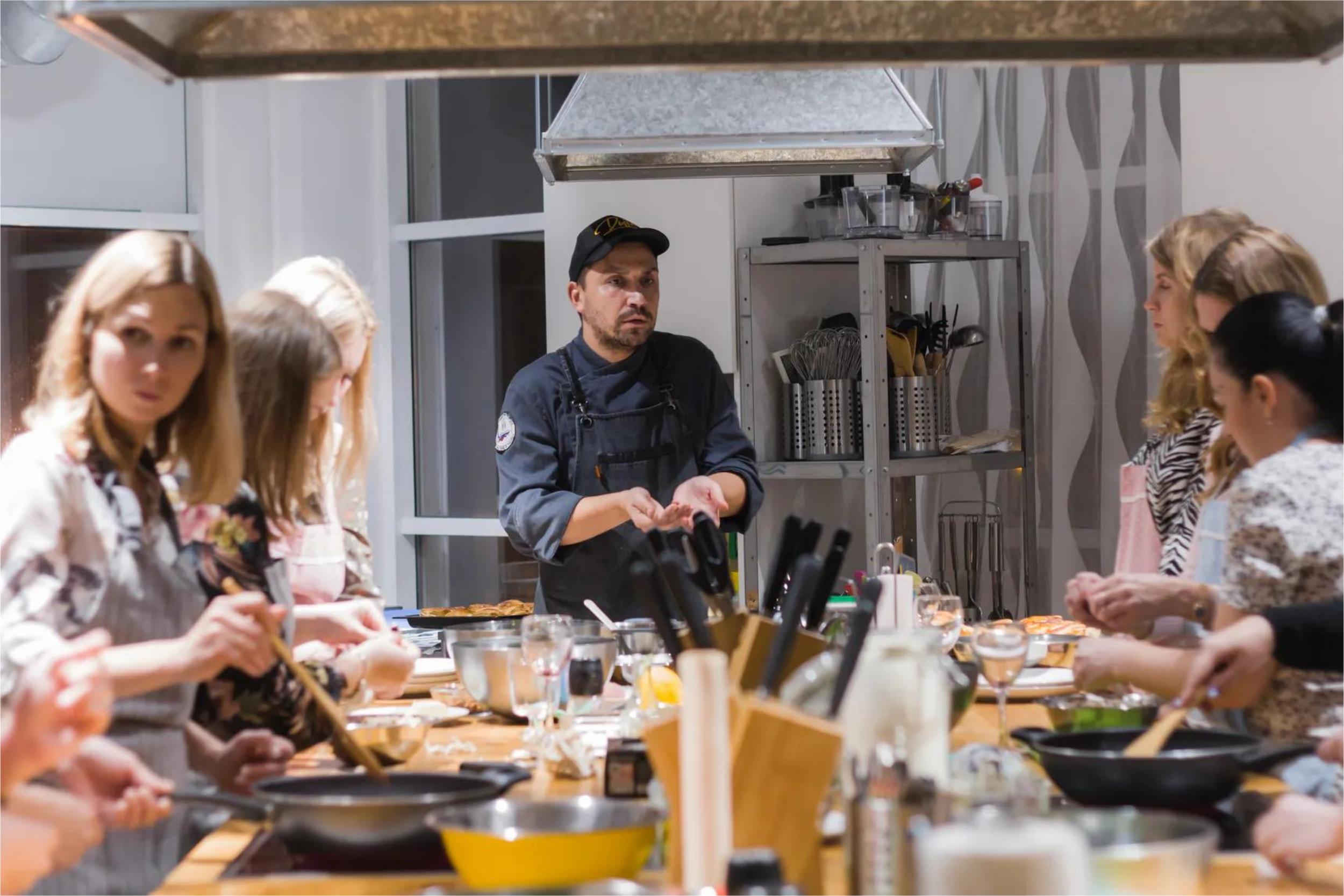 A cooking class with a chef instructor and students preparing food at a kitchen counter with various cooking utensils.