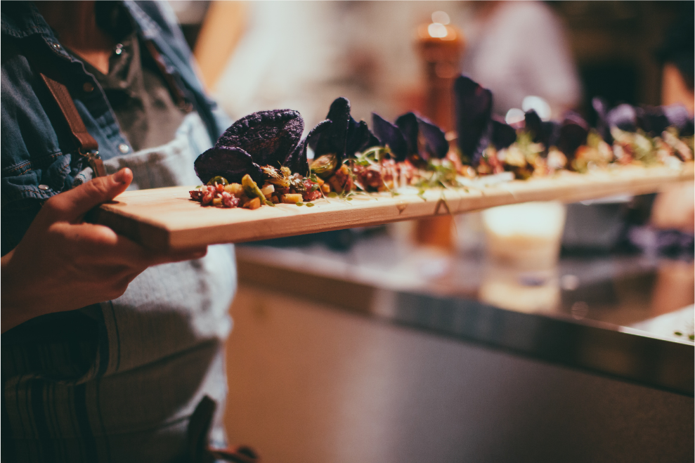 Person holding a wooden platter with colorful food, likely at a restaurant or event.