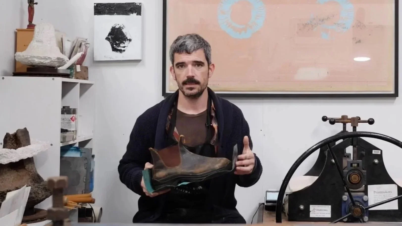 A man with gray hair and a beard holding a shoe in a workshop or studio, with shelves containing various items, artwork on the wall, and printing equipment visible in the background.