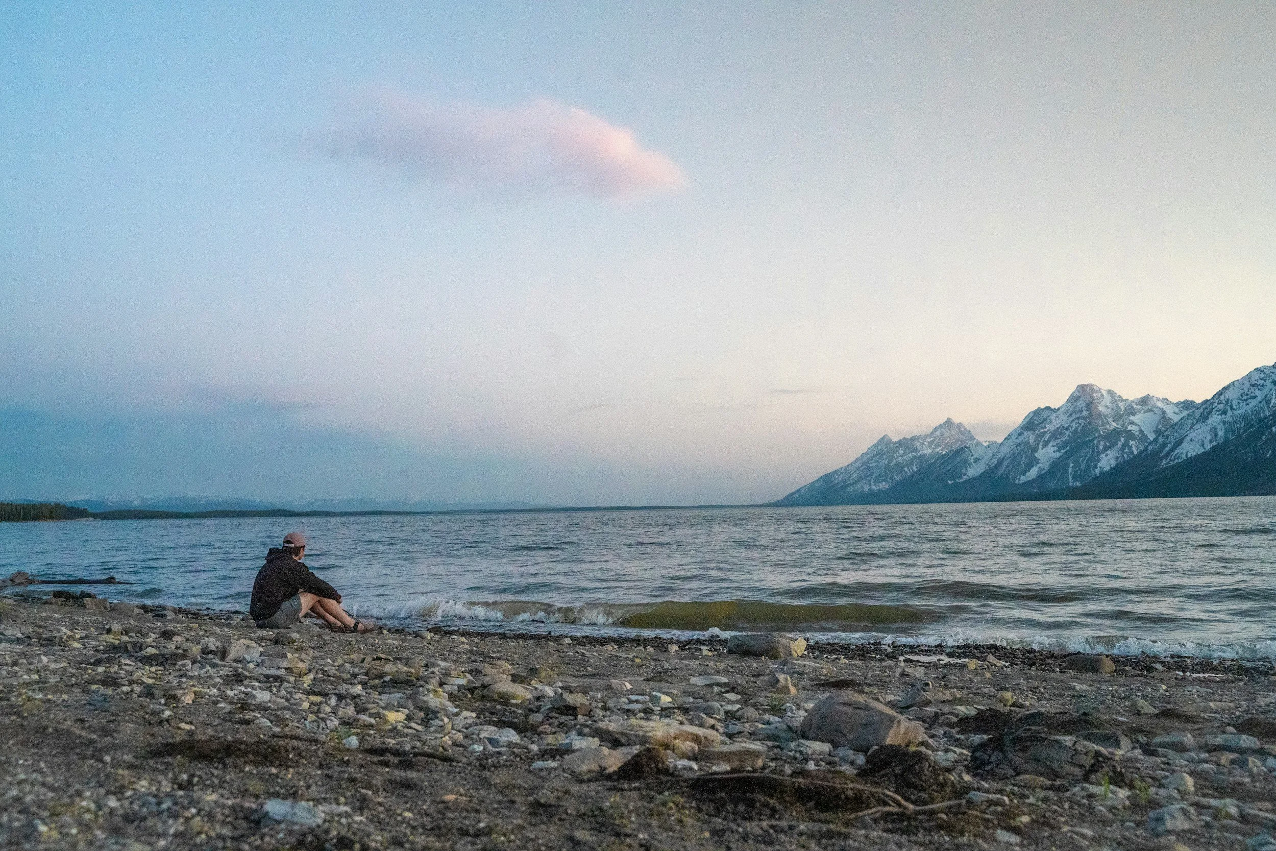 A man sits beside a lake in Wyoming at dusk, with the Grand Teton mountain range visible in the background.