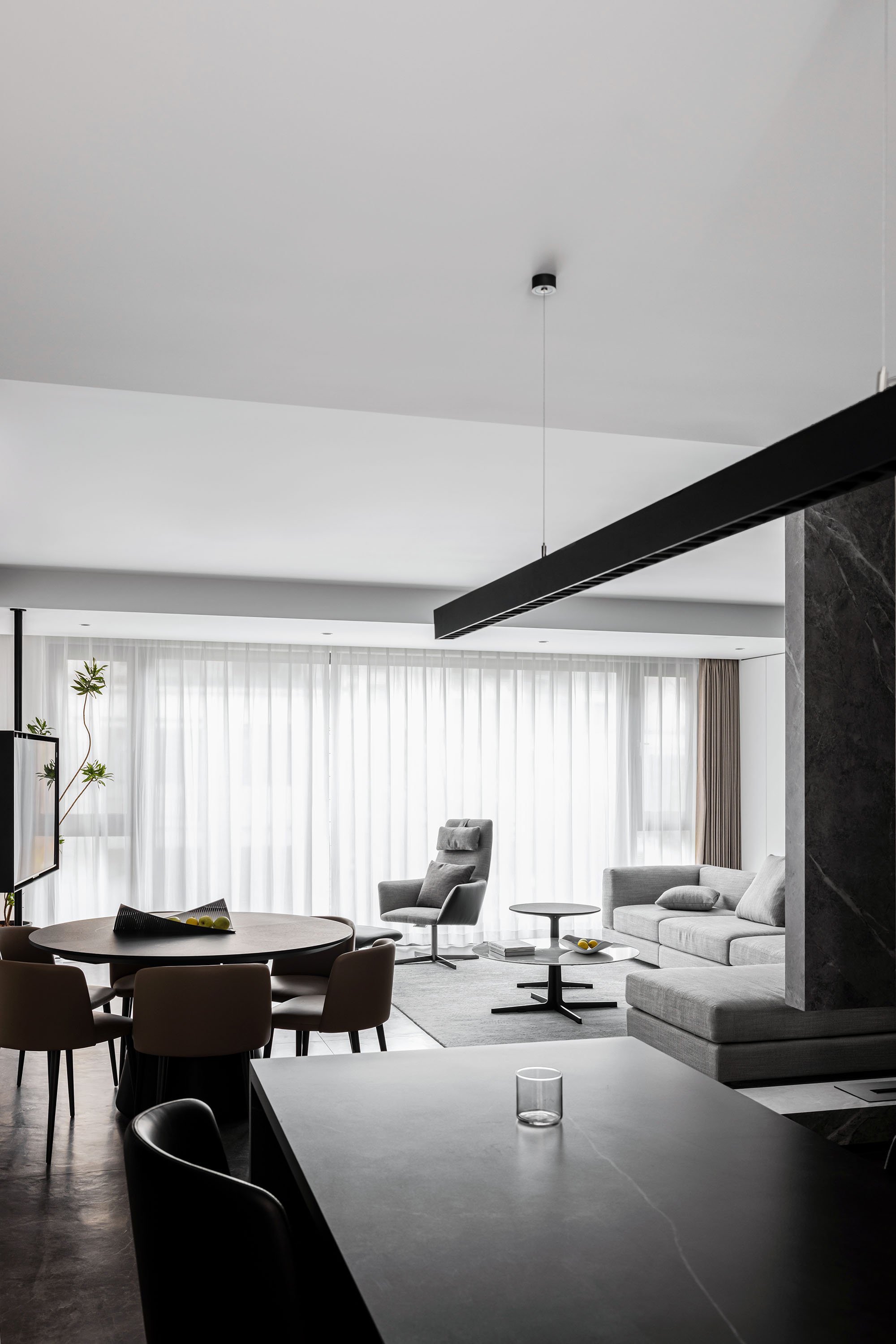 Modern living and dining area with a gray couch, armchair, round dining table, chairs, and large windows with sheer curtains, featuring a minimalist design.
