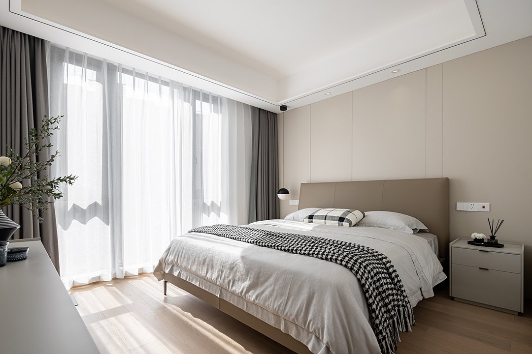 Modern bedroom with large window, king-size bed, checkered throw and pillow, bedside tables, and indoor plants.