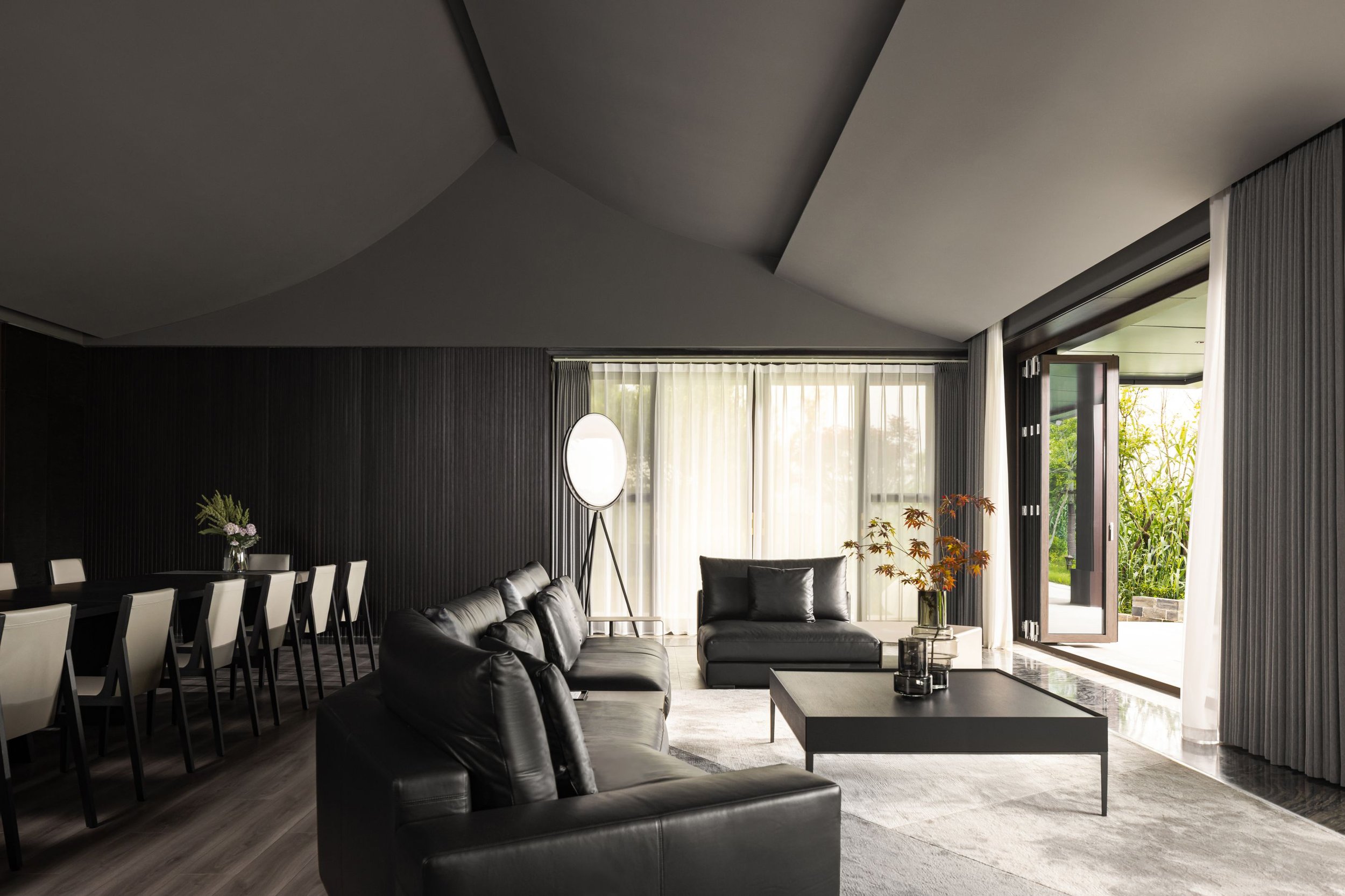 Modern living and dining room with black leather sofas, a long dining table with white chairs, a floor lamp, and a large window with sheer curtains. There is also a coffee table with decorative items and a view of greenery through an open door.
