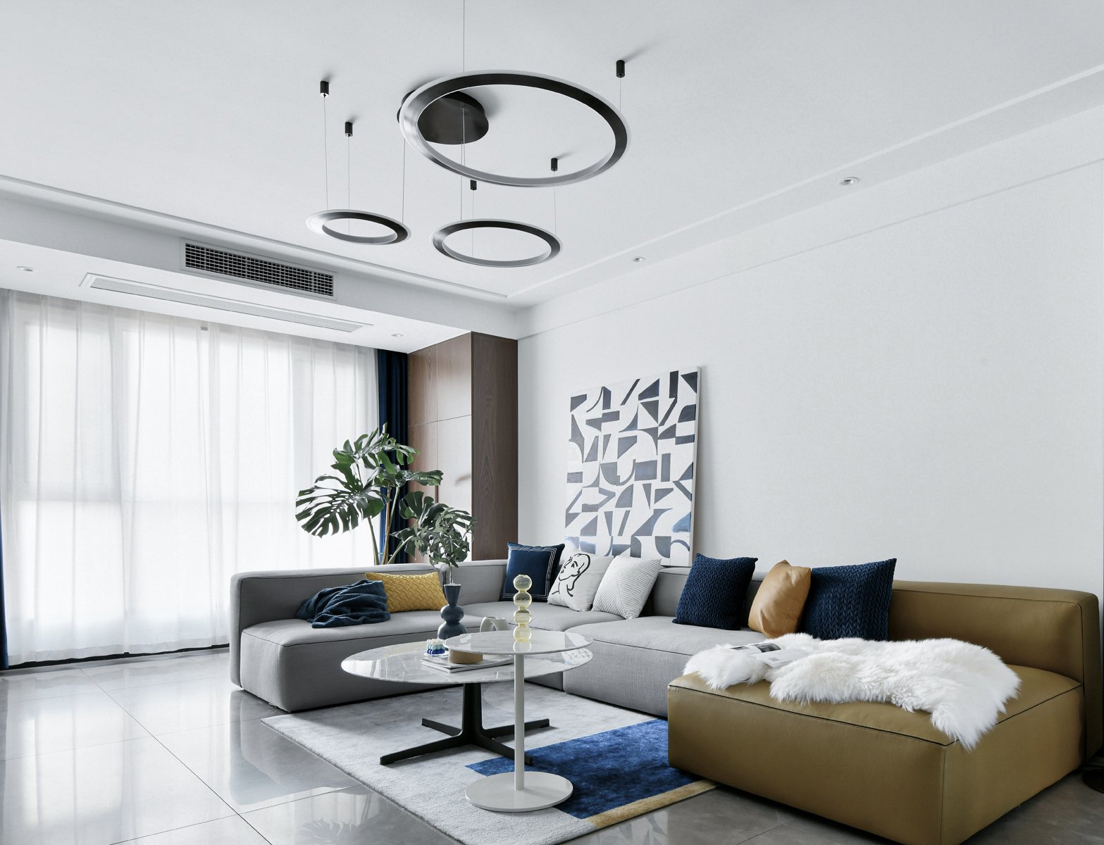 Modern living room with gray sectional sofa, geometric artwork, large plant, and minimalist chandelier. Features beige, blue, and yellow cushions, a marble coffee table, and white sheer curtains.