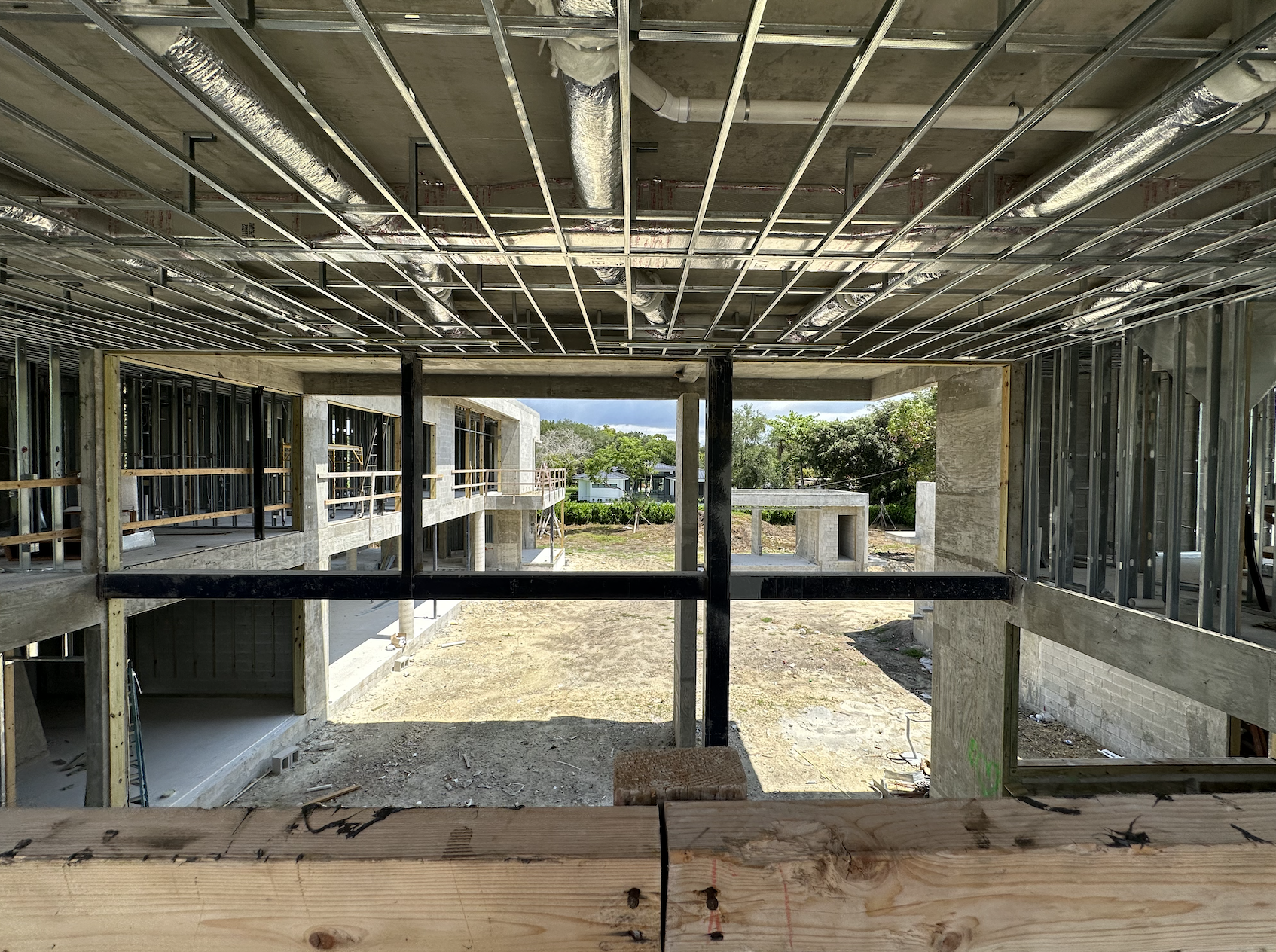 Construction site of a two-story building with exposed metal framework and ceiling ducts, overlooking an outdoor courtyard area.