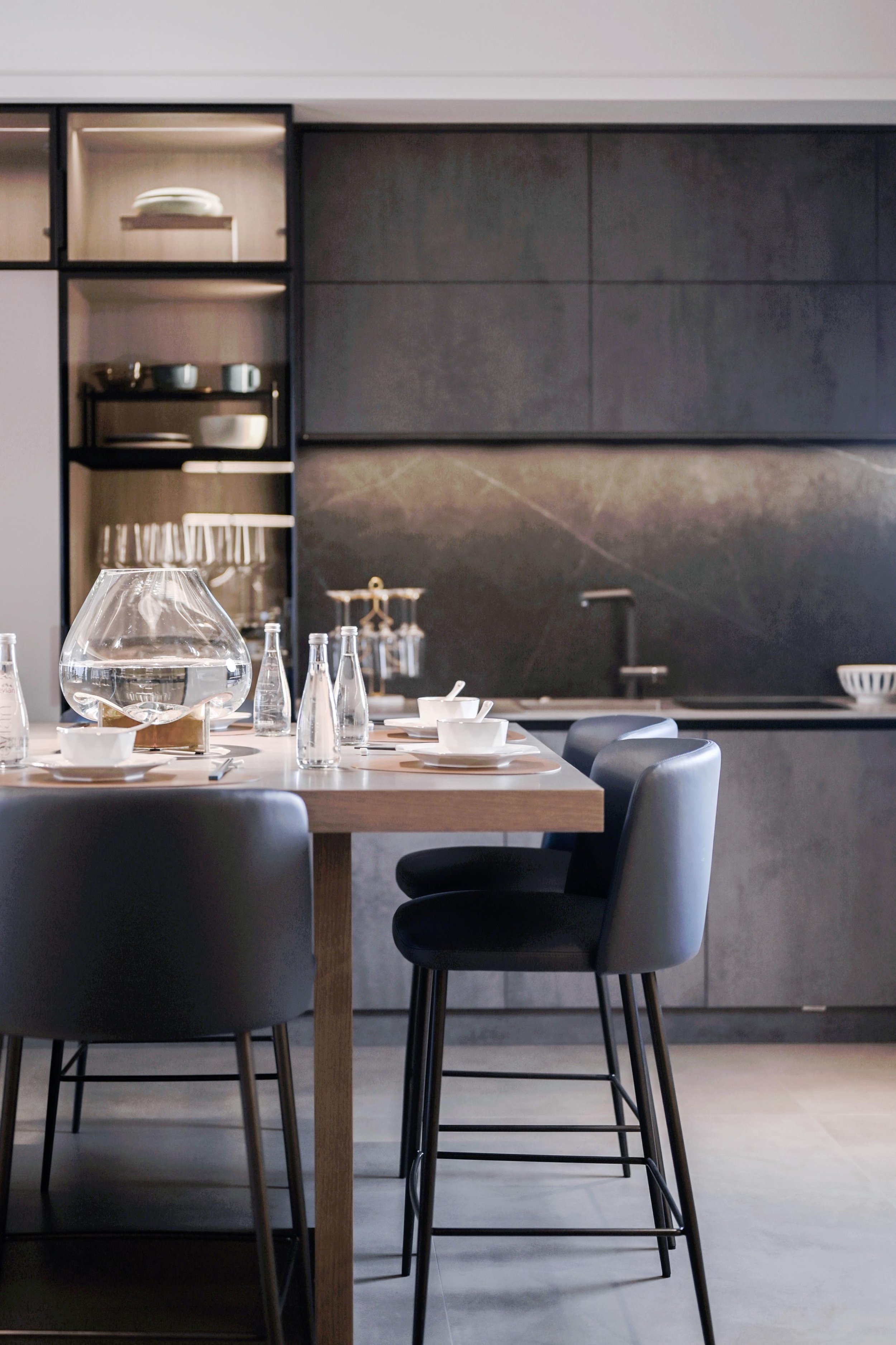 Modern kitchen with dark cabinetry, a dining table set with bowls, glasses, and water bottles, and high-back chairs.