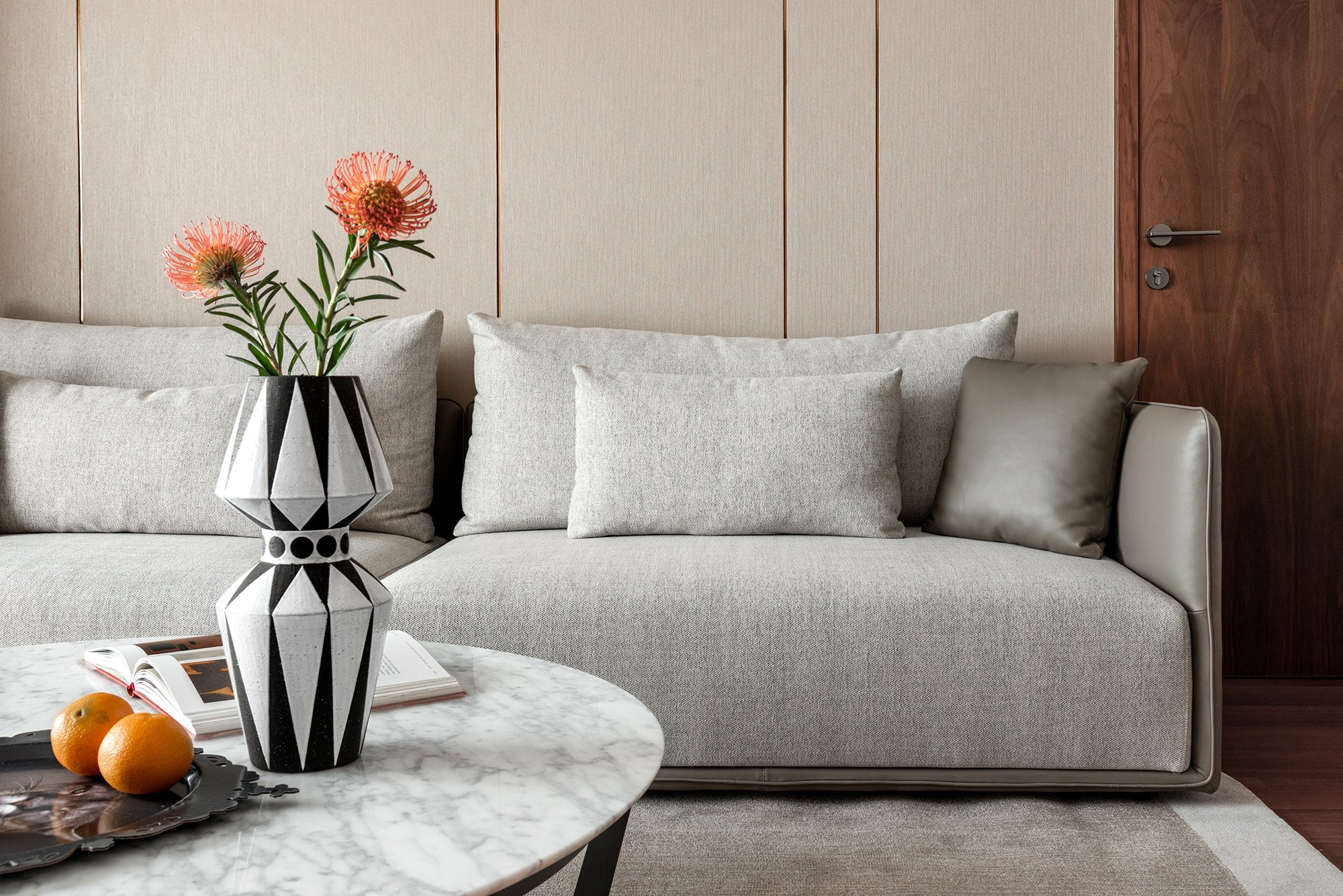 Modern living room with a gray sofa, beige cushions, marble coffee table with a vase of flowers and oranges, and a wooden door.