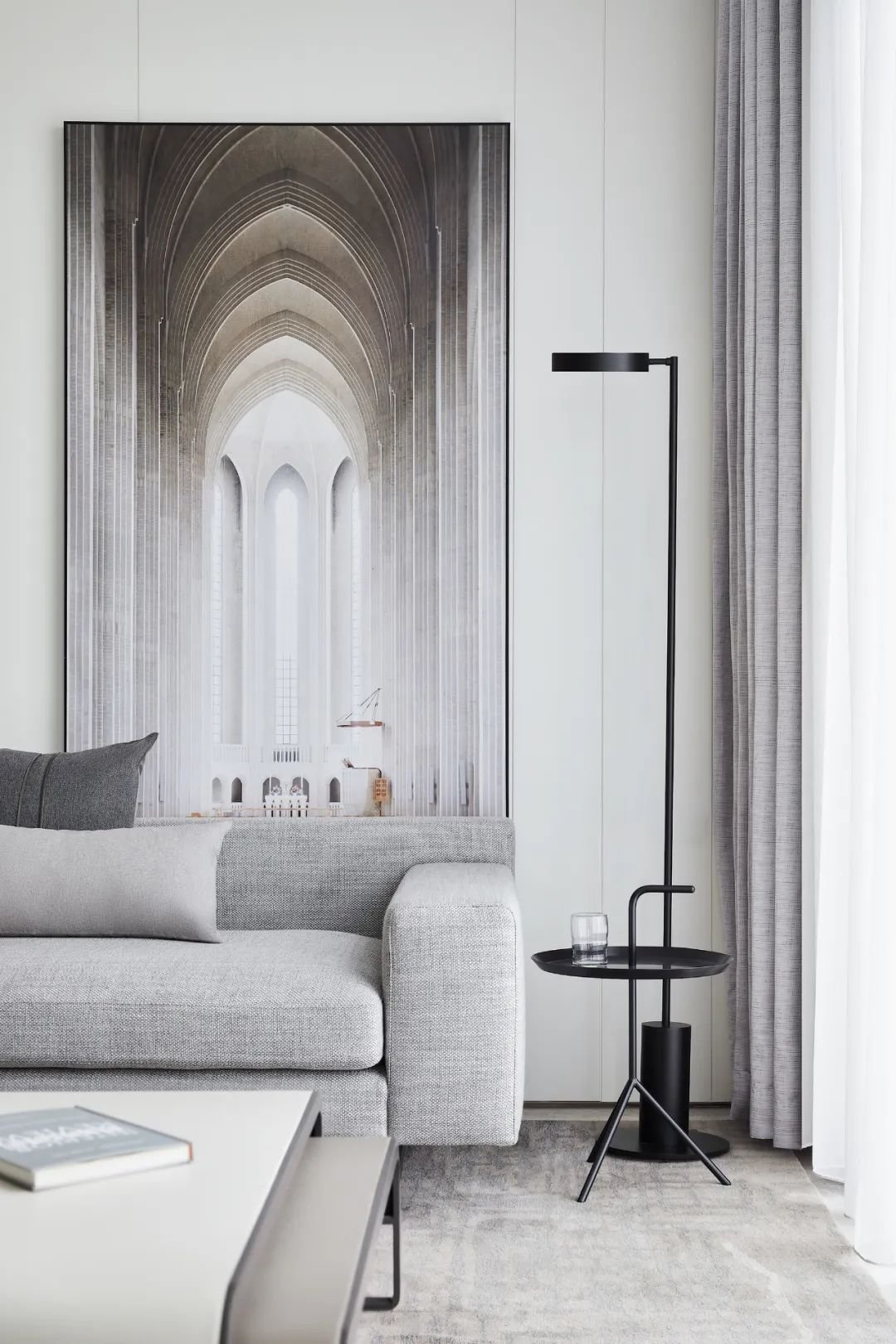 Modern living room with gray sofa, large architectural wall art, black floor lamp, small black side table with glass, and gray curtains.