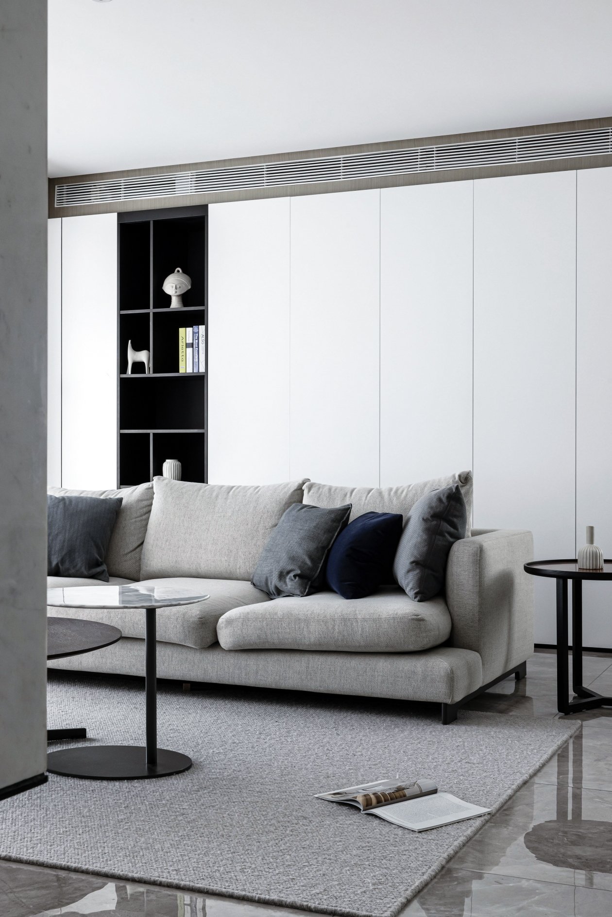 Modern living room with a gray sectional sofa, black coffee tables, and decorative pillows. A minimalist bookshelf with a few items and books is on the white wall. A magazine lies open on a gray rug.