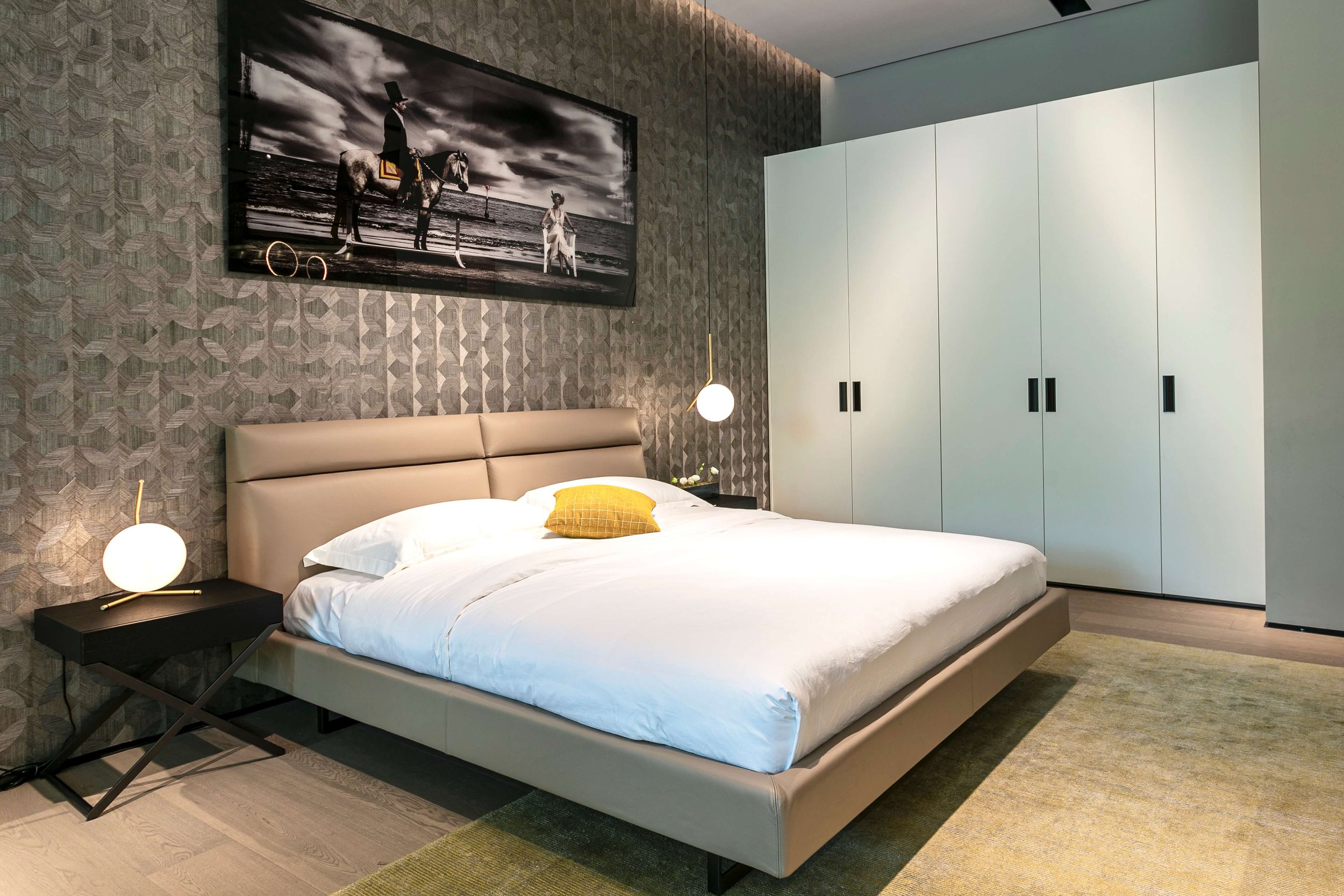 Modern bedroom with a beige platform bed, white bedding, and a yellow accent pillow. Nightstand with a spherical lamp. Large white wardrobe with black handles. Artistic black and white photograph on the geometric patterned wall. Ceiling-mounted penda