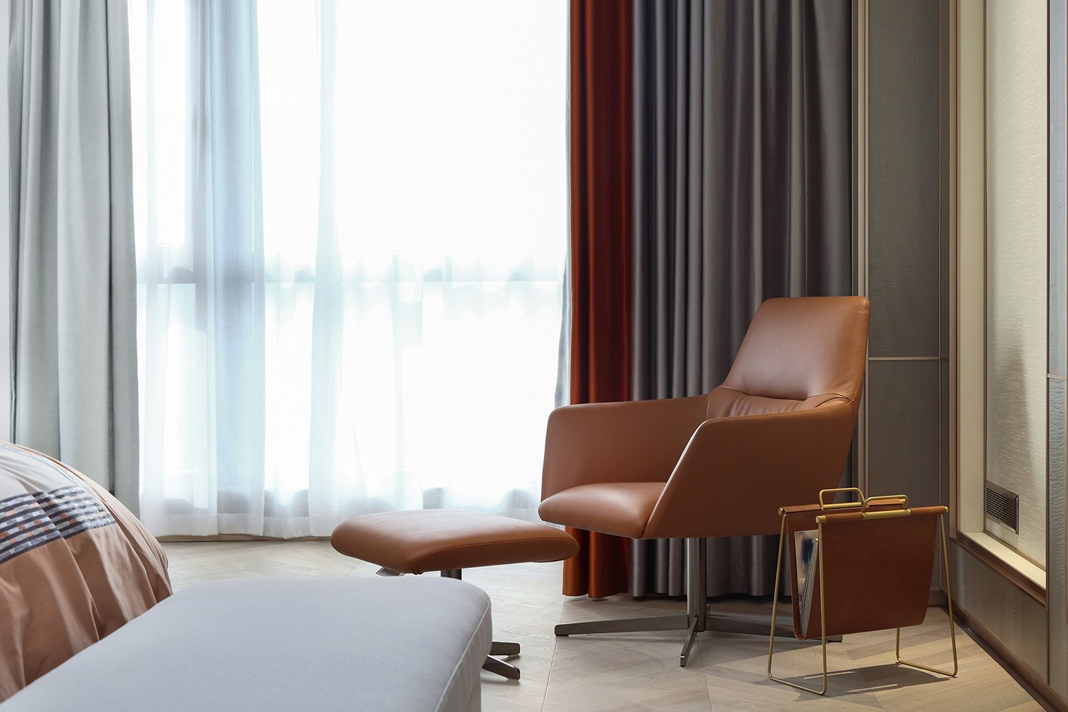 Modern lounge area with a brown leather armchair, matching footstool, and a magazine holder next to floor-to-ceiling curtains.