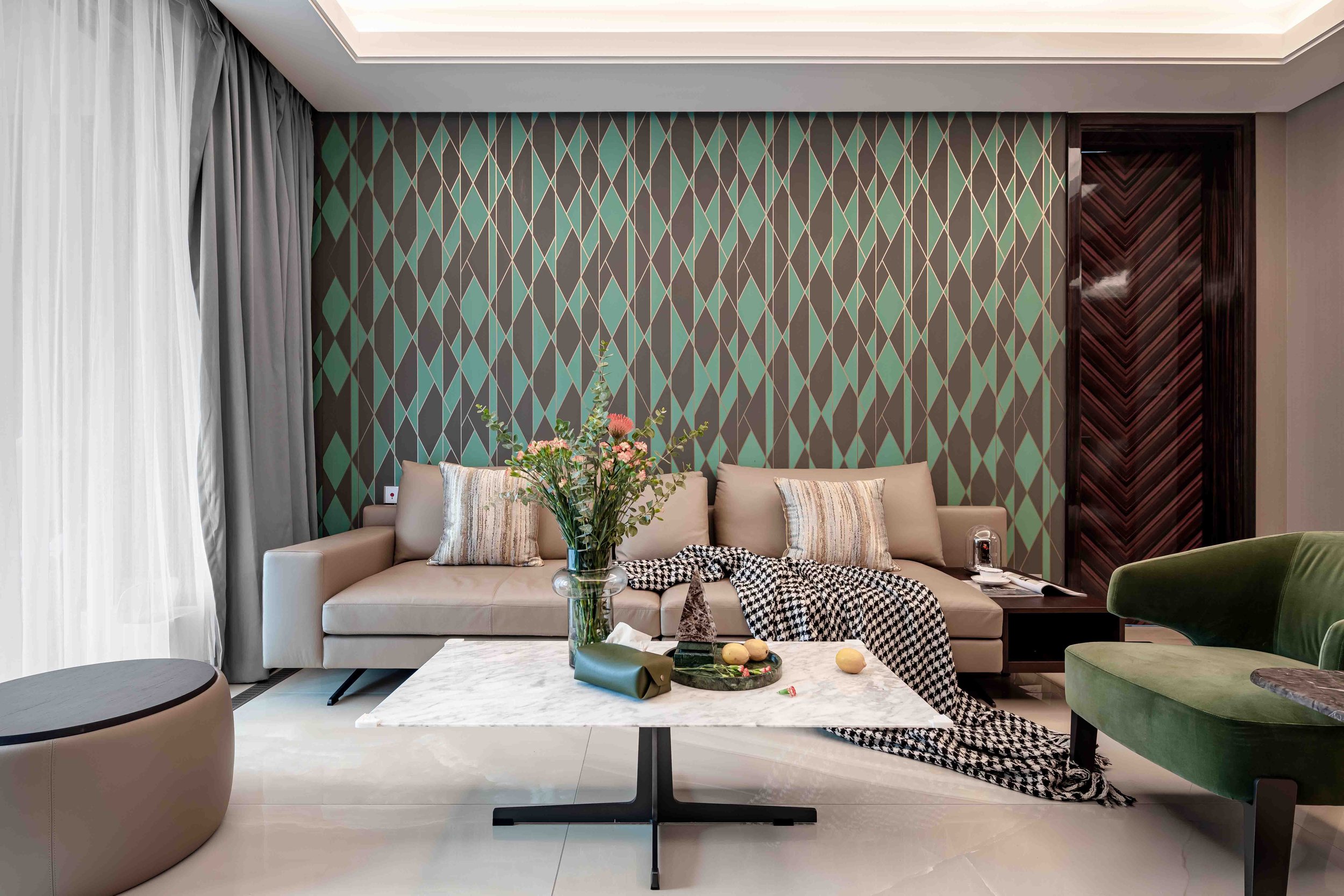 Modern living room with a beige sofa, green patterned wall, and a marble coffee table. There is a green chair, a vase with flowers, decorative pillows, and a checkered throw blanket on the sofa. Large windows with light gray curtains.