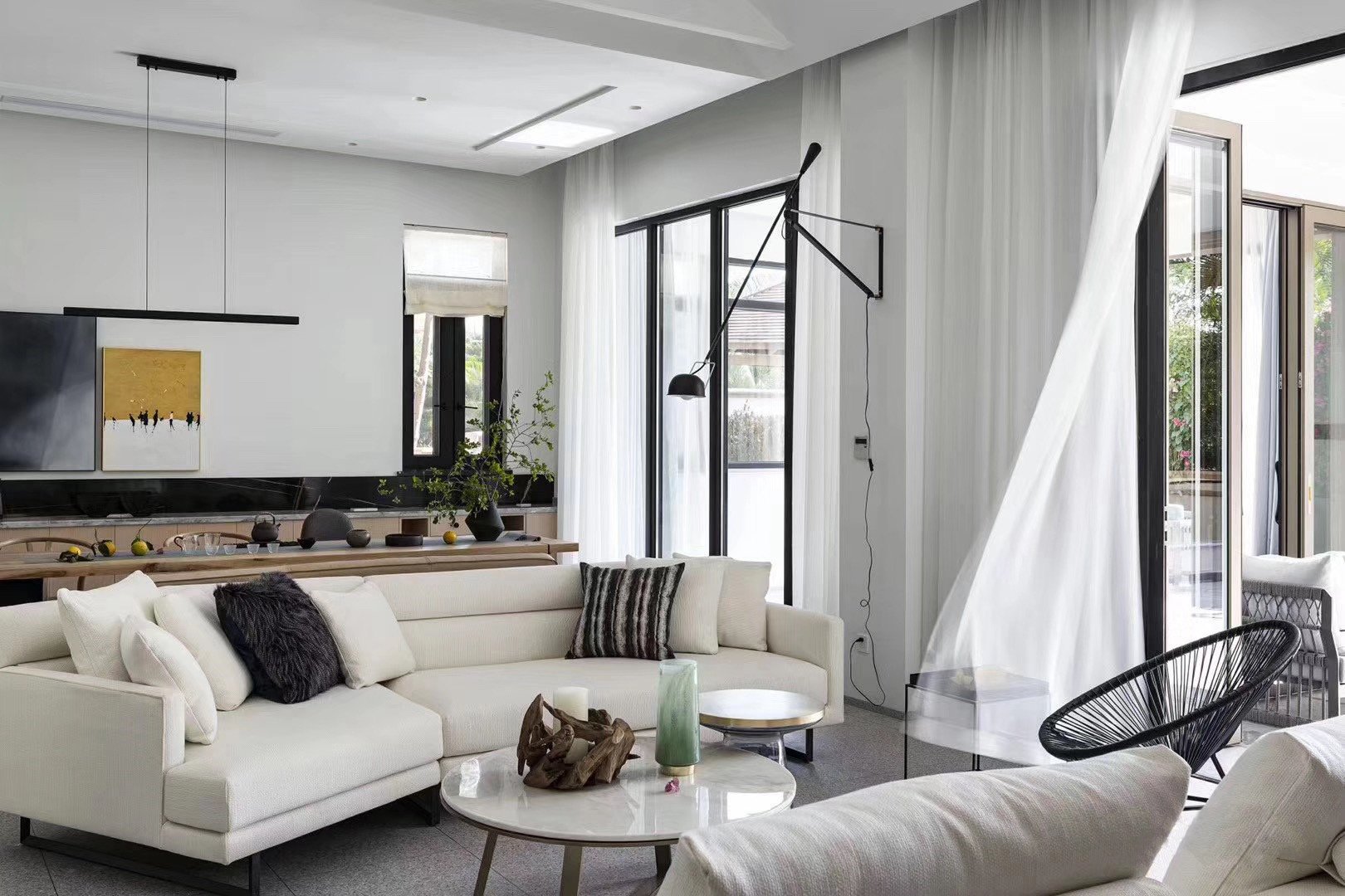 Modern living room with white sectional sofa, black metal and glass accent tables, and large windows with sheer curtains. Decorative plants and artwork on wall.
