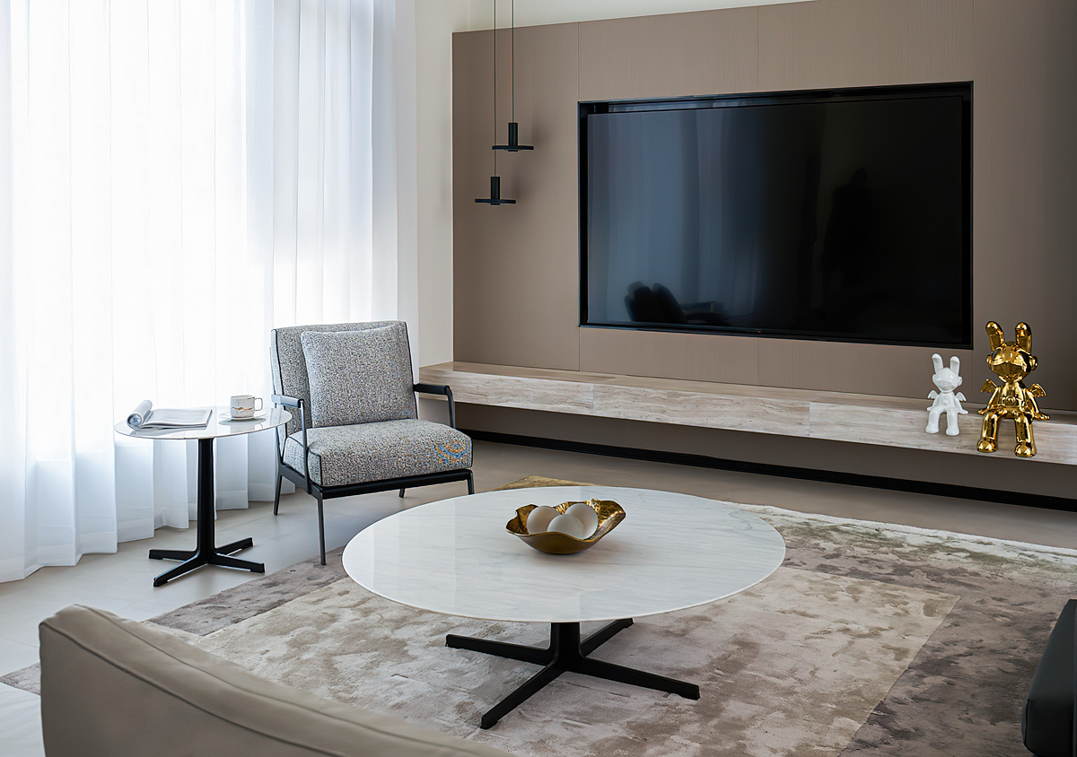 Modern living room with large TV on wall, marble coffee table, gray armchair, side table with a cup and magazine, gold and white decorative bunnies, sheer curtains, and beige rug.