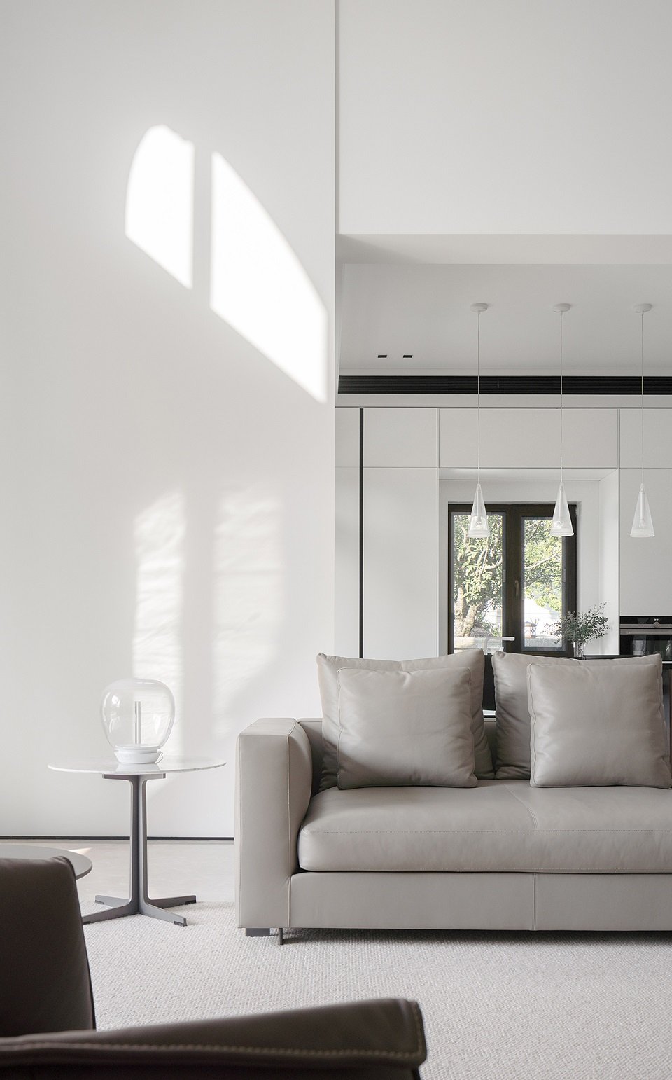 Modern living room with gray sofa, pillows, white walls, round side table with a glass vase, pendant lights, large window, and sunlight casting shadows.