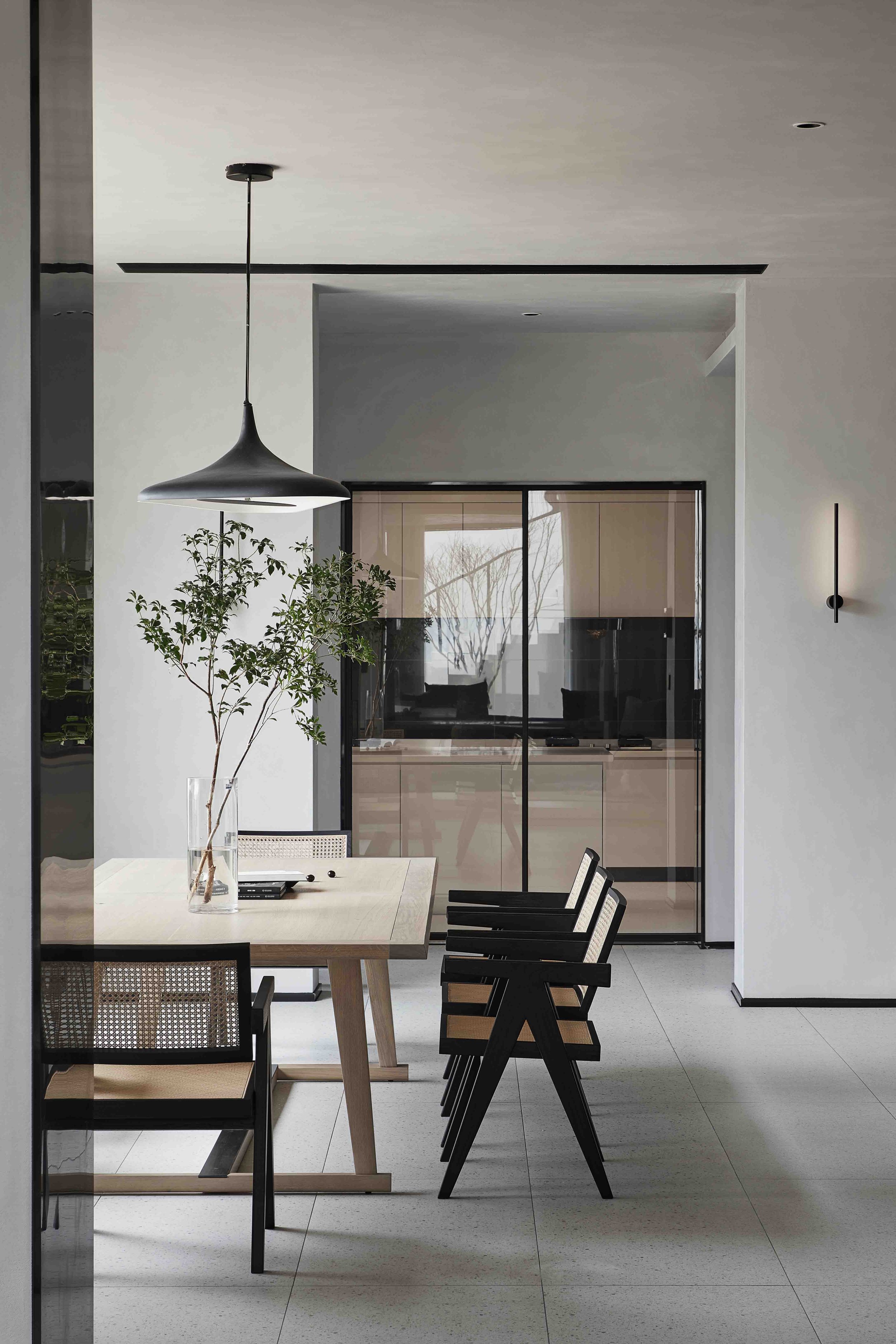 Modern dining room with wooden table, black chairs, minimalist pendant light, and glass vase with branches, leading to a kitchen with glass doors.