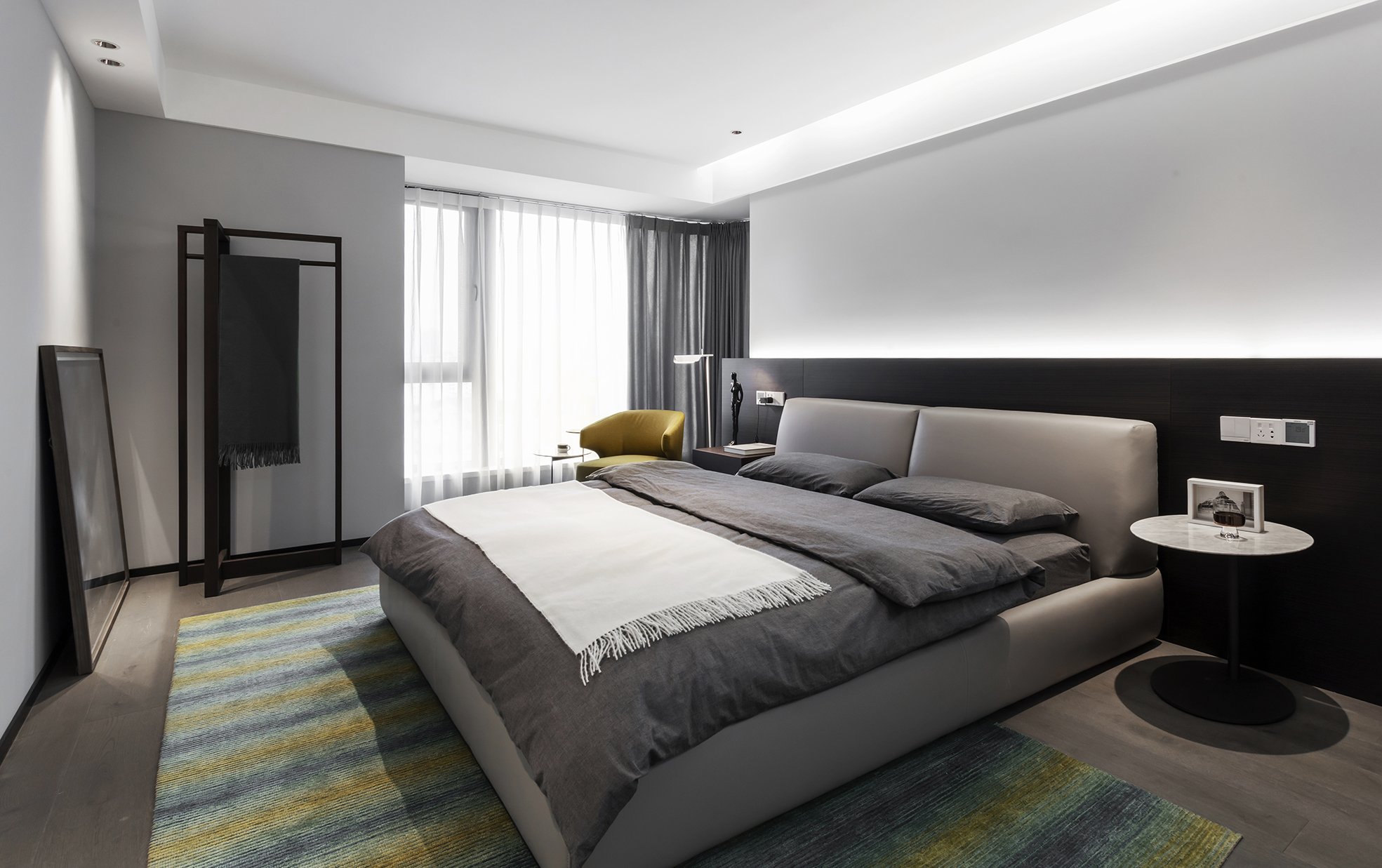 Modern bedroom interior with a gray bed, gray and white bedding, a small round side table, a fabric chair, and a striped area rug. The room features floor-to-ceiling curtains and minimal decor.
