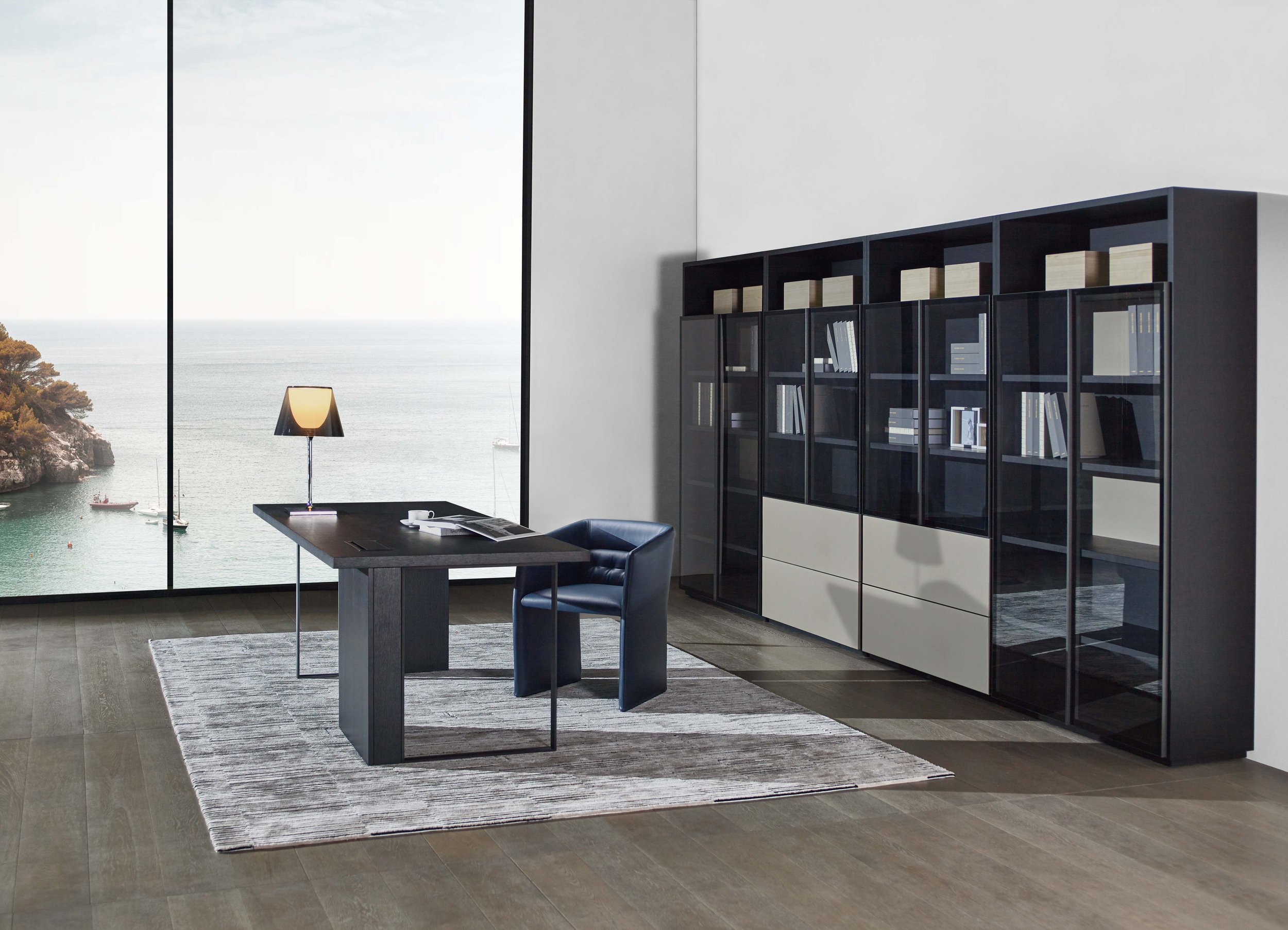 Modern office interior with a large window, a dark desk with a lamp and papers, a blue chair, and a bookshelf, overlooking a scenic ocean view.